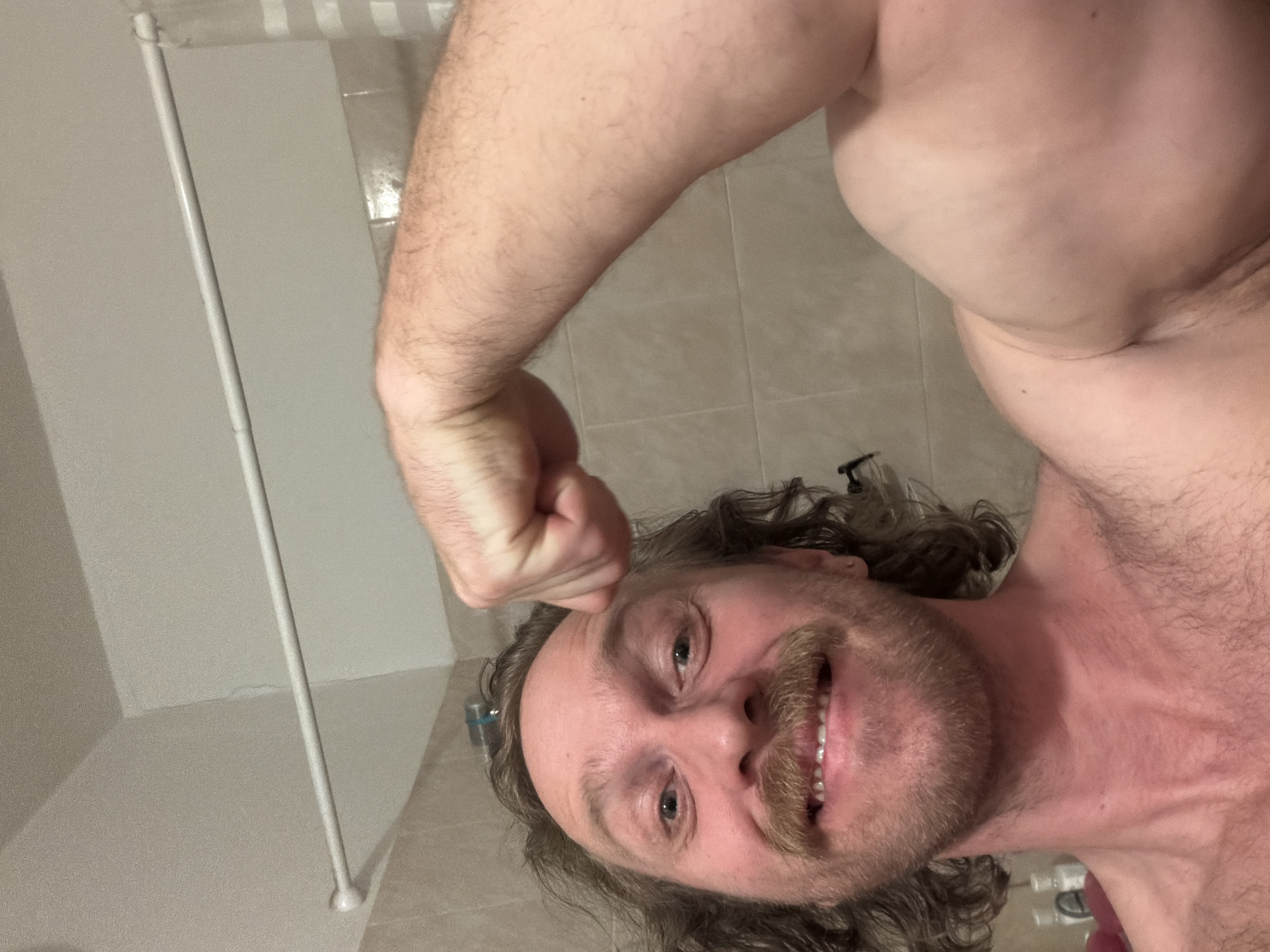 A man with long, wavy hair and a large, handlebar mustache poses with his right arm raised and fist clenched, showcasing a flexed bicep. He is smiling at the camera. The setting appears to be a bathroom, with light-colored tiles visible in the background and a white shower curtain rod in the lower left of the frame. The scene does not provide any information to locate the image within the city of Saint Petersburg, Russia.