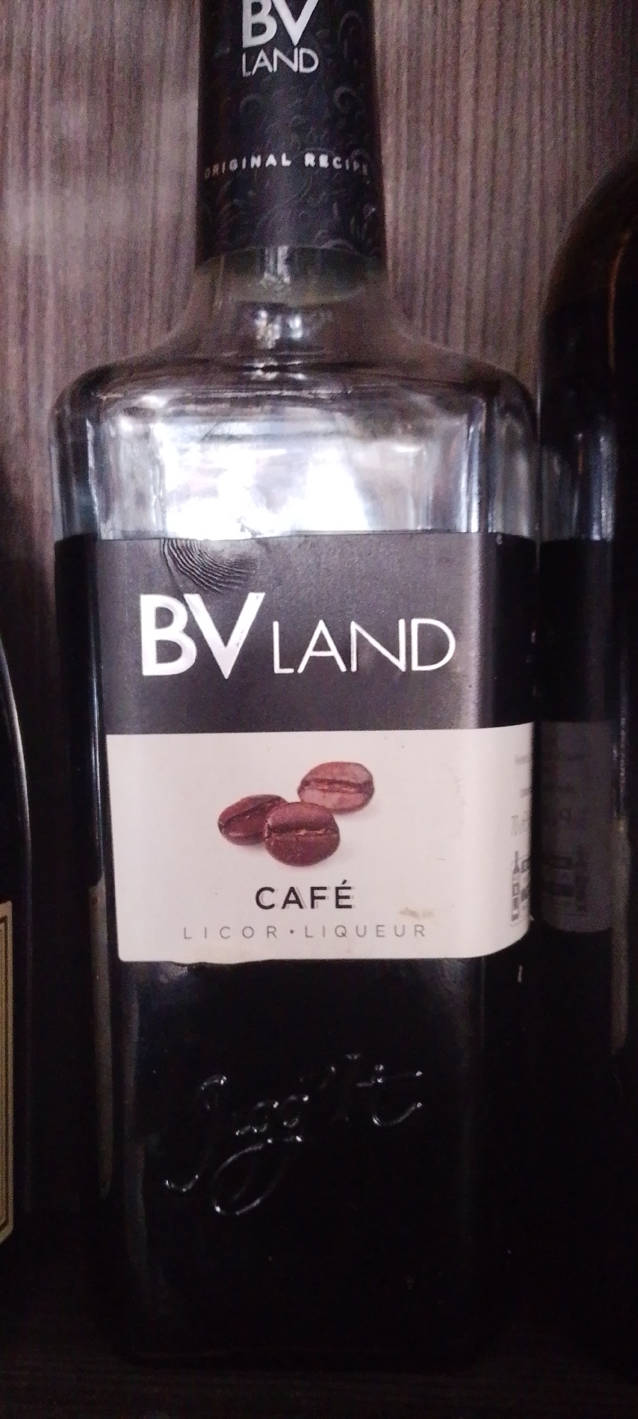 The image shows a close-up of a glass bottle of "BV LAND CAFÉ LICOR LIQUEUR". The bottle has a black label with white lettering. The label features three coffee beans as a graphic element above the text "CAFÉ" and "LICOR. LIQUEUR". Below the label, the bottle's glass is embossed with a cursive word, possibly a brand name. The bottle appears to be on a shelf or surface with a wooden texture in the background. There is no information in the image to suggest the location is in the city of Howayi, Nigeria.