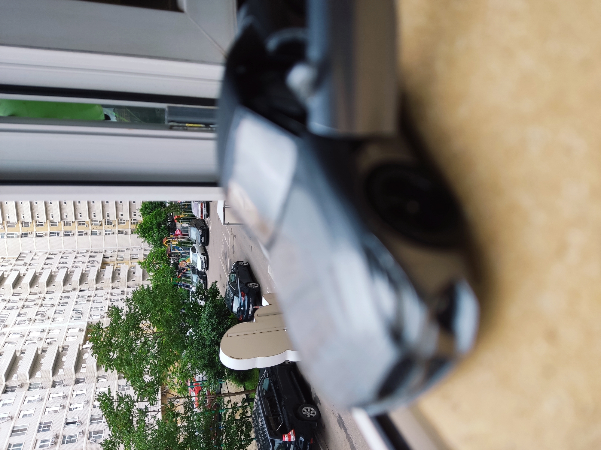 The image shows a view from a window, likely of an apartment building overlooking a courtyard. The location is indicated to be Krasnodar, Russia.
Visible outside the window are several parked cars in the courtyard, green trees, and a playground with colorful equipment. In the background, a large, multi-story residential building with multiple balconies and windows is seen. The building appears to be densely populated.
The window frame is visible in the foreground, partially obscured by an out-of-focus object (likely a household appliance or device). The window appears to be partially open.
The overall scene suggests a typical urban residential area in Krasnodar. There are no people immediately visible in the frame.