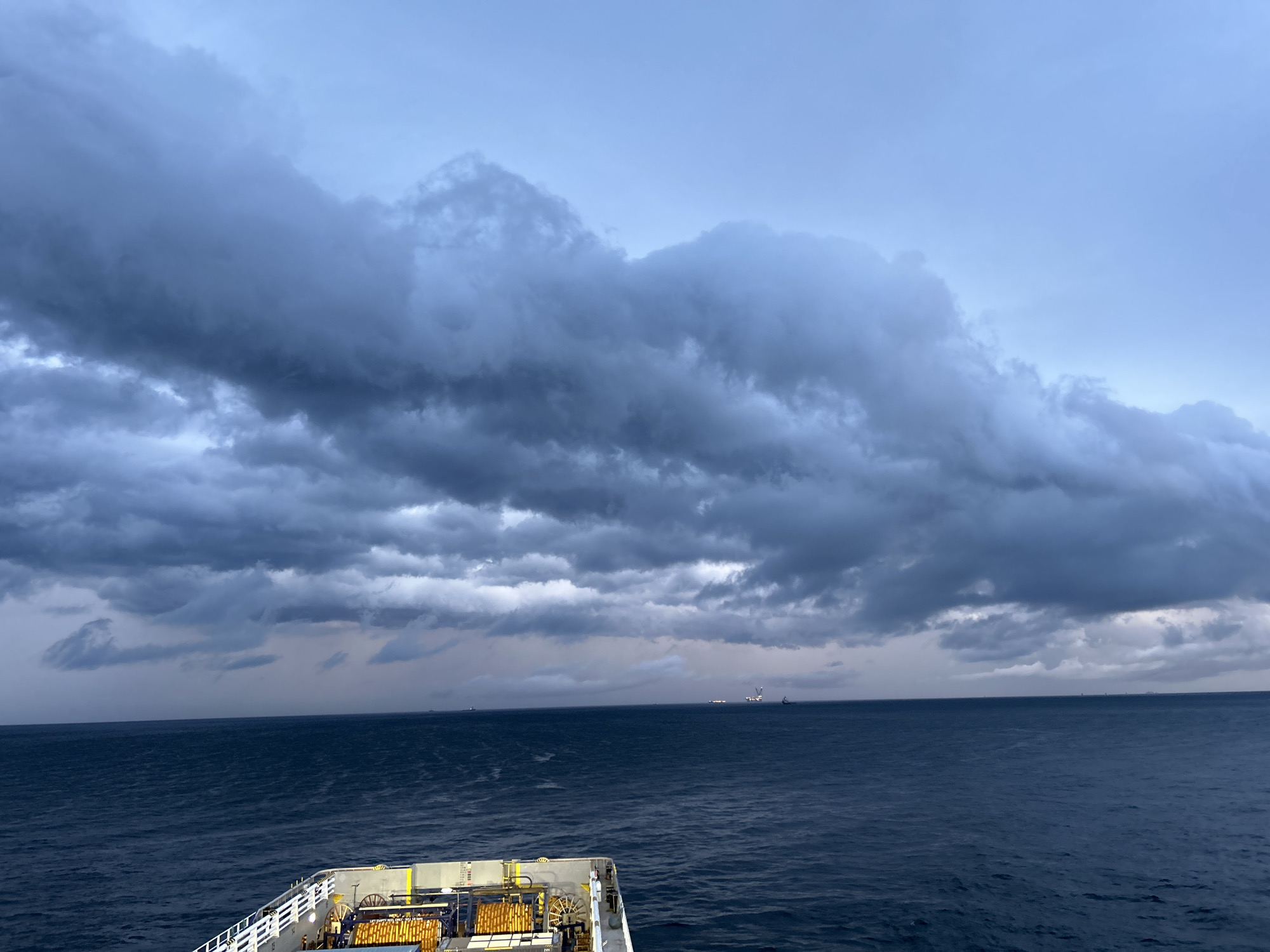 The image presents a dramatic seascape viewed from the deck of a large vessel, likely a ship or ferry. The scene is dominated by a vast, dark, and cloudy sky that stretches across the entire upper portion of the frame. The clouds are heavy, dark grey, and textured, suggesting overcast or potentially stormy weather. Above these dense clouds, the sky lightens to a muted, pale blue-grey, especially towards the upper right.
The horizon line separates the turbulent sky from the deep blue-grey ocean, which occupies the middle ground. The sea surface has visible ripples and small waves, indicating movement, but it appears generally calm despite the ominous sky. In the far distance on the horizon, several indistinct structures are visible. To the middle-right, a more prominent, multi-level structure with vertical elements, possibly an offshore platform or a large industrial vessel, can be discerned. Other smaller, darker shapes are faintly visible to the left and far right on the distant horizon.
In the foreground, the structure of the vessel's deck is clearly visible, occupying the bottom quarter of the image. It features white railings, grey metal surfaces, and yellow machinery or equipment. Coiled cables or ropes are stacked on the deck, and various structural components are visible. On one of the yellow structures, some text is legible, including "LIFT EMPTY HOOK ONLY" and "MAX 1.0T", indicating operational instructions or weight limits. Another marking "SWL MAX 2.0T" is visible on yellow equipment further right on the deck.
The time of day appears to be either very early morning, late afternoon, or simply a heavily overcast day, as the light is subdued and diffused, creating a somber and moody atmosphere. The overall impression is one of a journey across a vast, open sea under powerful, dramatic weather conditions.