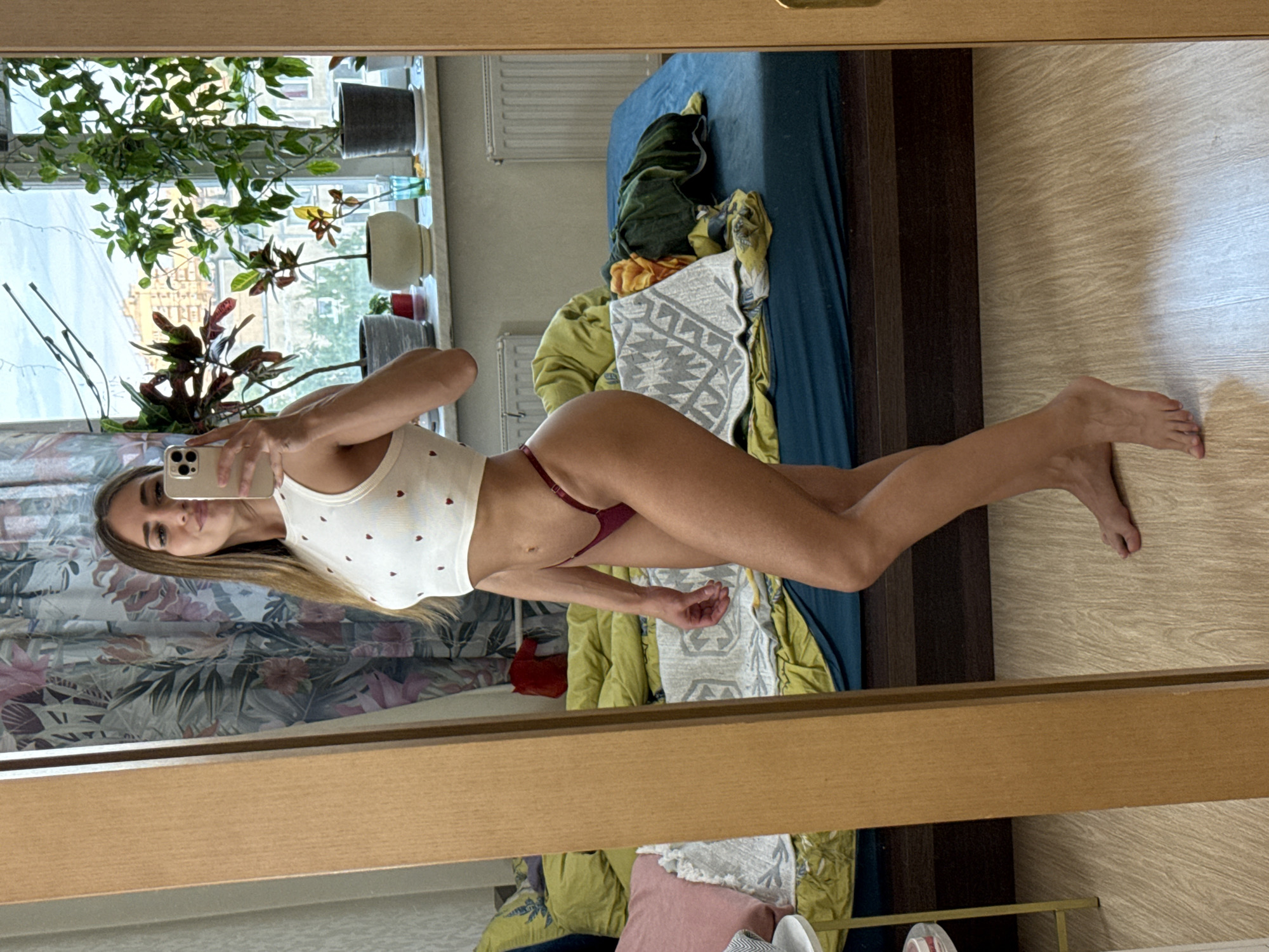 A young woman with long, blonde hair is taking a selfie in a mirror. She is wearing a white crop top with small red hearts and burgundy thong underwear. She is standing in what appears to be a bedroom, with a bed in the background covered in blankets and pillows. There is a window with plants on the sill behind her, and curtains hanging on the wall. The room has light-colored wooden floors and a wooden-framed mirror. The woman is looking at her phone, which she is holding in her right hand. The scene is lit by natural light coming from the window. The location is identified as Unknown, Russia.
