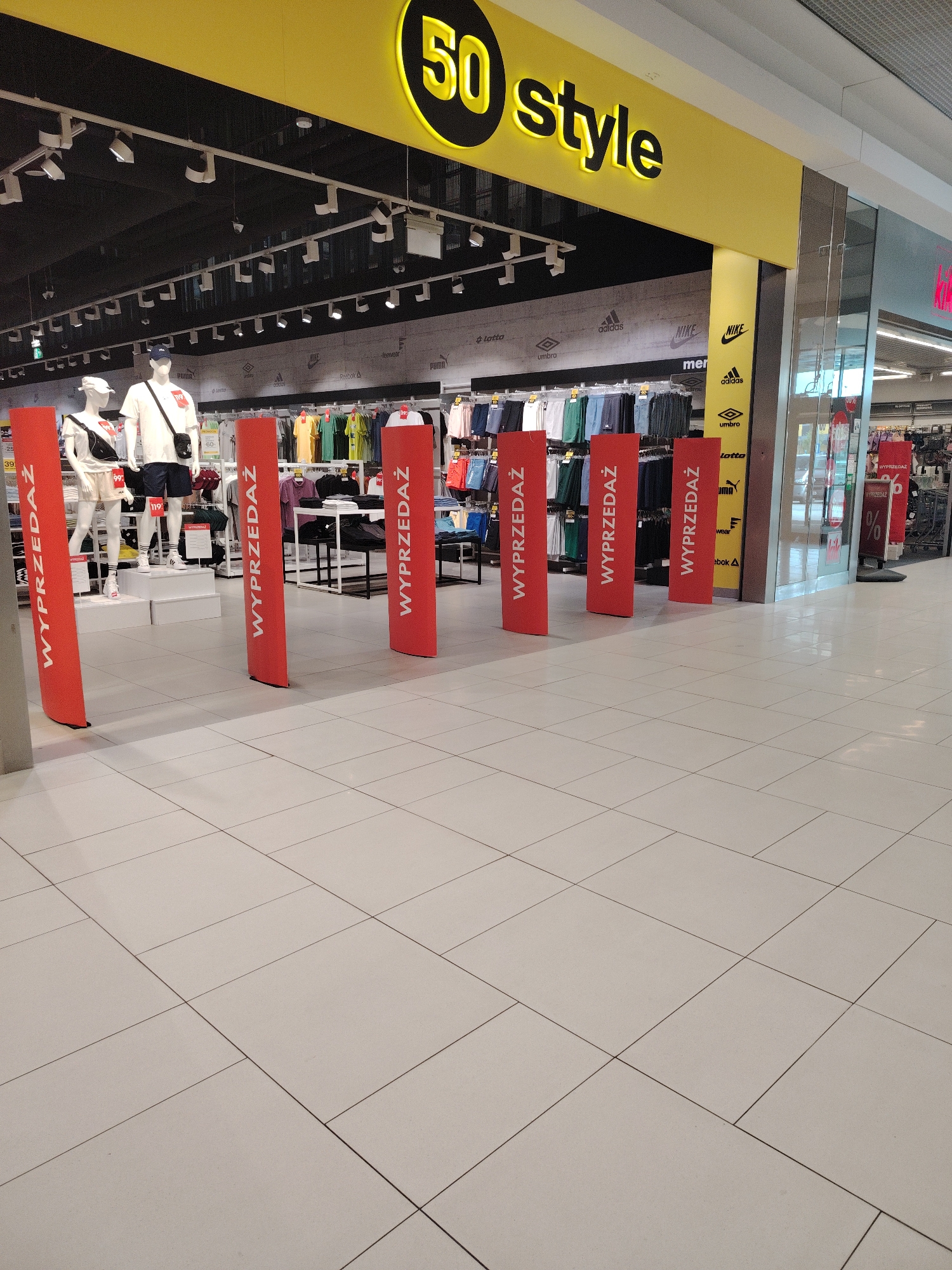 The image shows the entrance to a "50 Style" store in Wołomin, Poland, identified by the store's prominent yellow sign with black lettering. The store appears to be a clothing or shoe retailer, as evidenced by mannequins displaying outfits and racks of clothing visible inside. The entrance is partially blocked by several tall, red vertical banners with white text that reads "WYPRZEDAŻ," meaning "SALE" in Polish. These banners are spaced out, creating an aisle leading into the store. Inside the store, clothing is displayed on racks, and there are lighting fixtures on the ceiling. The floor in front of the store is tiled with large, light-colored square tiles.