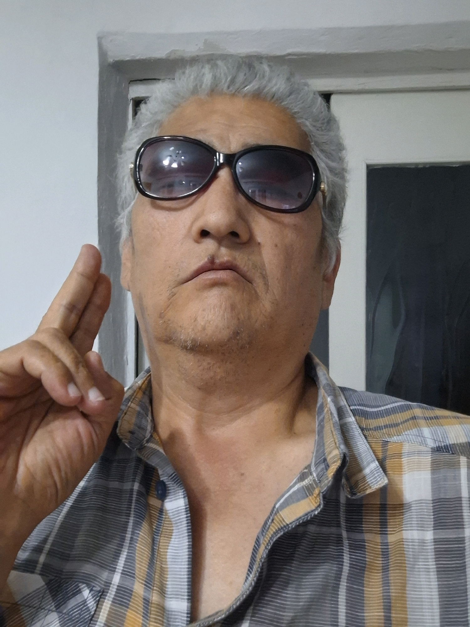 A medium shot captures an adult male with graying hair and sunglasses indoors. He is wearing a plaid shirt and has his left hand raised in a gesture with two fingers extended. The background consists of a plain wall and a doorway, suggesting an interior setting. No specific location in Almaty, Kazakhstan, can be determined from the image.
