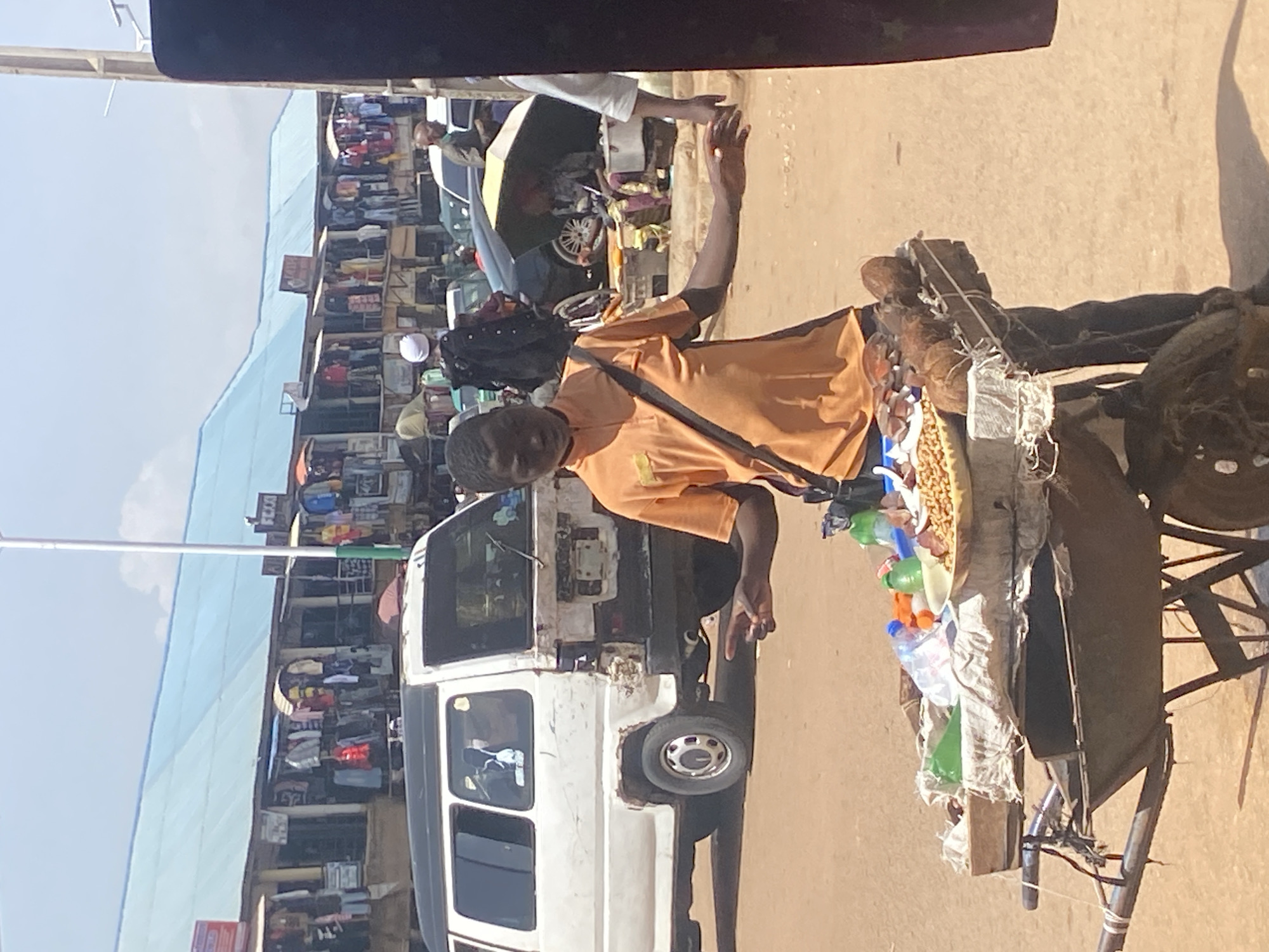 A young boy stands behind a wheelbarrow filled with food items and coconuts in Bukuru, Nigeria. He wears an orange polo shirt and dark pants, with a bag slung across his chest. His left hand is raised, fingers splayed, as if gesturing or making a point, while his right hand is extended towards the viewer. The wheelbarrow contains a bowl of what appears to be nuts or seeds, several bottles of water, and two coconuts. In the background, a white van is parked, and beyond that, a row of market stalls with blue roofing is visible, displaying clothing and other goods. Several cars and other people are present in the scene, suggesting a busy market environment. The ground is dirt, and the sky is clear with some clouds.