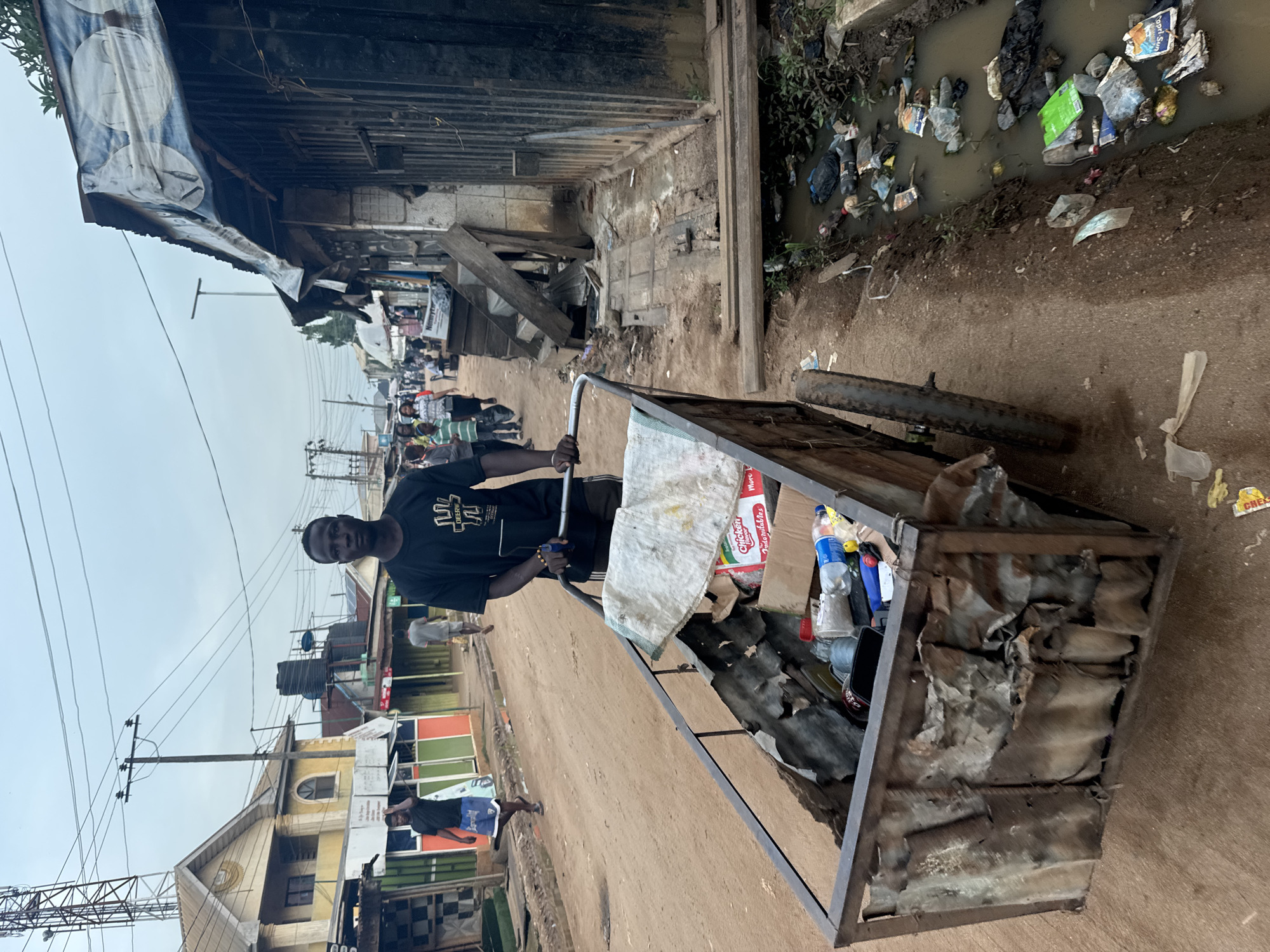 The image, rotated 90 degrees clockwise, captures a street scene in Ihiagwa, Nigeria, under an overcast sky during daytime. A young man, positioned slightly left of center, stands holding the handlebars of a two-wheeled, homemade metal cart. He wears a black short-sleeved t-shirt with a white graphic design that includes the word "DEBBY" and dark trousers. He looks directly towards the viewer with a calm expression.
The cart, made of a metal frame and corrugated materials, is filled with various items, including plastic bottles, a white cloth, and a "Chicken Indomie" noodle packet. One of the cart's wheels is visible on the right side, resting on the unpaved, reddish-brown dirt road.
To the right of the man and the cart, an open drainage ditch runs parallel to the road. This ditch is filled with murky water and a significant amount of discarded plastic waste and debris, including a "Capri-Sun" pouch and other packaging.
The background reveals a bustling streetscape. On the left, a series of makeshift shops or stalls line the road, with one structure featuring a blue banner adorned with two large white circular patterns. Further down the street, several pedestrians are visible. Utility poles carrying numerous electrical wires crisscross the sky. In the mid-ground, a light yellow building with an arched window stands out amongst other residential or commercial structures. The scene depicts a lively, informal urban or semi-urban area with characteristic infrastructure and evident waste management concerns.