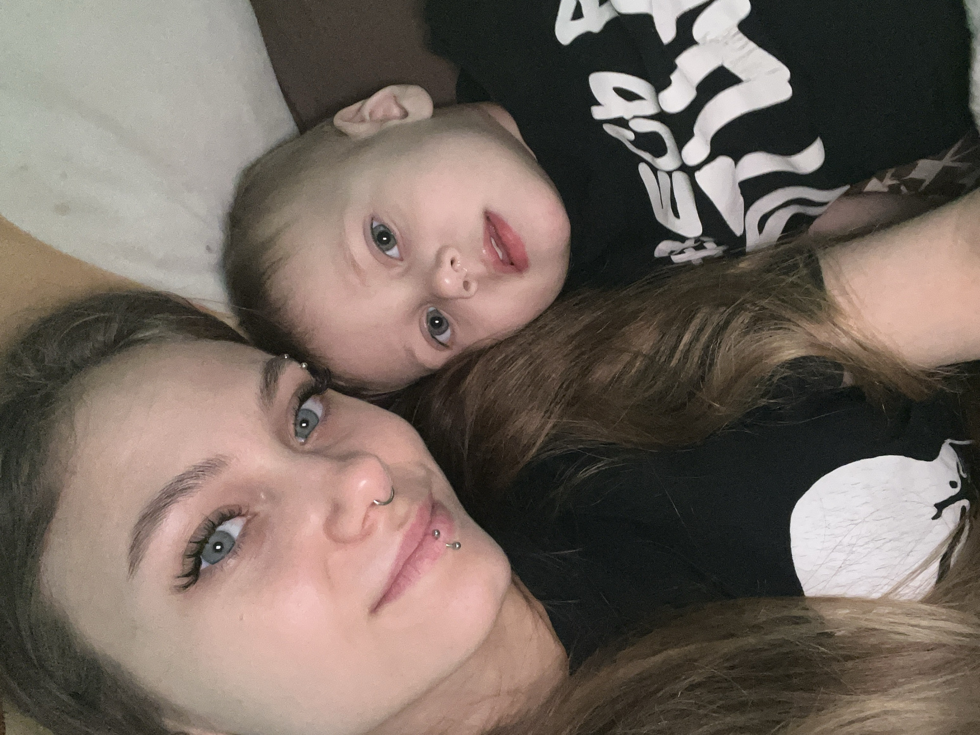 A selfie-style image shows a woman and a baby lying down together. The woman is in the foreground, looking at the camera with light blue eyes and brown hair. She has piercings in her eyebrow, nose, and lip. The baby is lying behind her head, also looking at the camera with blue eyes. The baby is wearing a black shirt with a white skeleton design. The woman is wearing a black shirt with a white design on the shoulder. The background is a mix of light and dark colors, possibly a couch or bedding. The location is not discernible from the image.