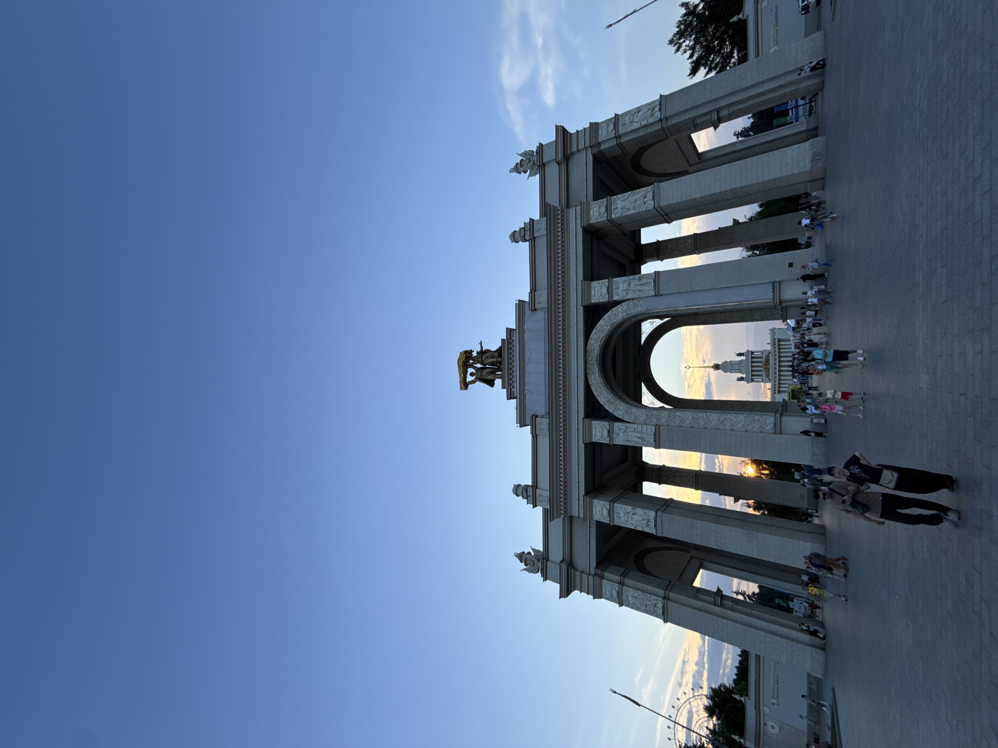 The image shows the Main Arch of VDNKh (Exhibition of Achievements of National Economy) in Moscow, Russia, during sunset. The monumental arch, constructed from concrete and adorned with sculptures, stands prominently in the foreground, its grand arches framing a distant view of a building with a spire. The sky above is a clear, deep blue, with a few wispy clouds near the horizon where the sun is setting, casting a warm glow. A crowd of people is visible in the plaza in front of the arch, some walking, others standing. A Ferris wheel is visible in the far left background. The paving of the plaza is made of dark gray rectangular stones. The overall scene captures a public space with significant architectural features under a fading sky.