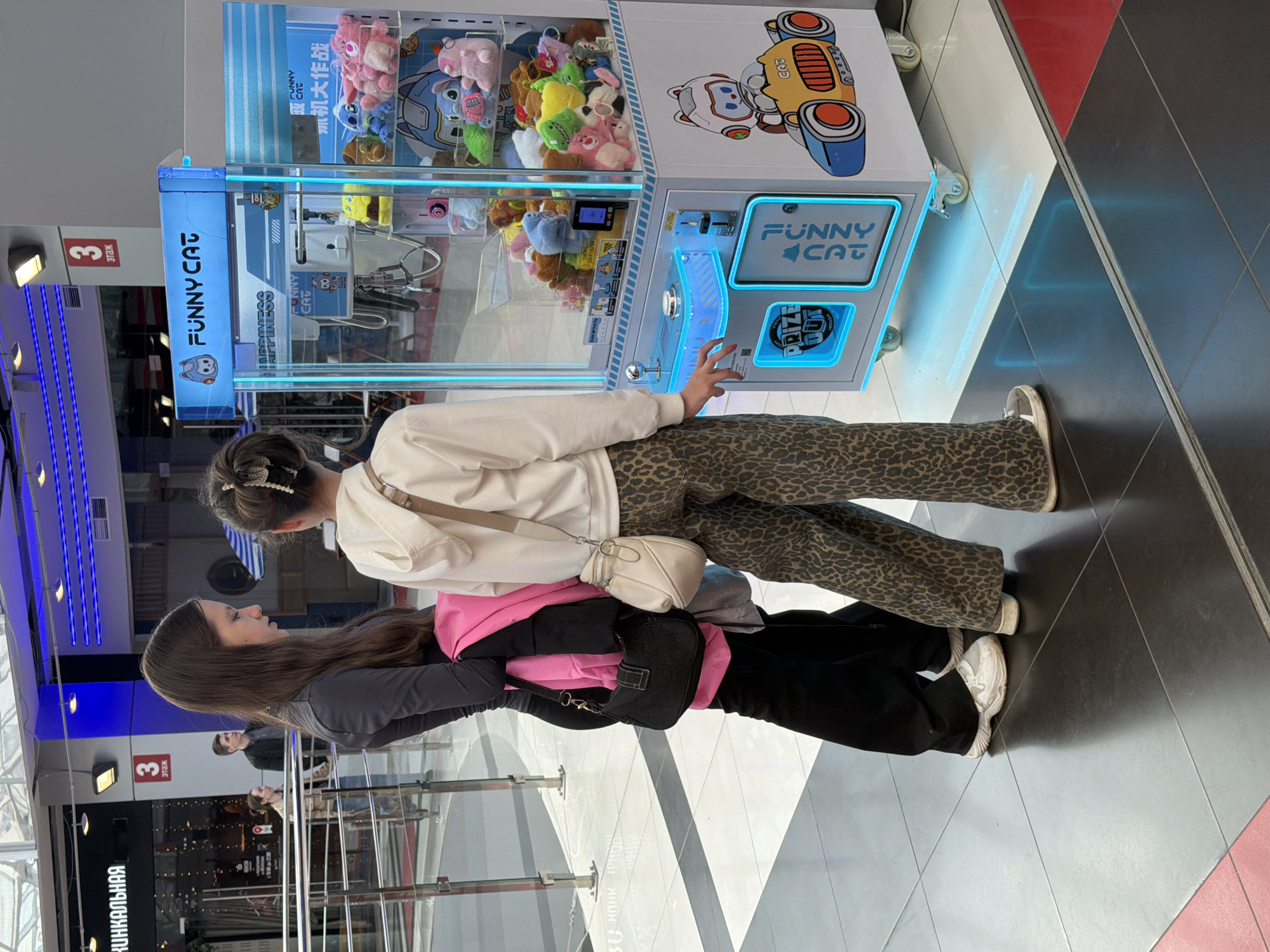 Two women are standing in front of a "Funny Cat" claw machine filled with plush toys, likely attempting to play it. The woman closest to the machine is reaching towards the machine with her right hand. She is wearing a white sweater, leopard print pants, and a light-colored crossbody bag. The other woman is behind her, wearing a grey jacket, a pink garment underneath, and black pants. A black purse hangs from her shoulder. They are in a shopping mall or arcade with a tiled floor and glass barriers. Signs in Russian are visible in the background. The sign says “КИНКАЛЬНАЯ”. Another sign says “3 Этаж”, which means floor 3.
The image was taken in the city of Izhevsk, Russia.