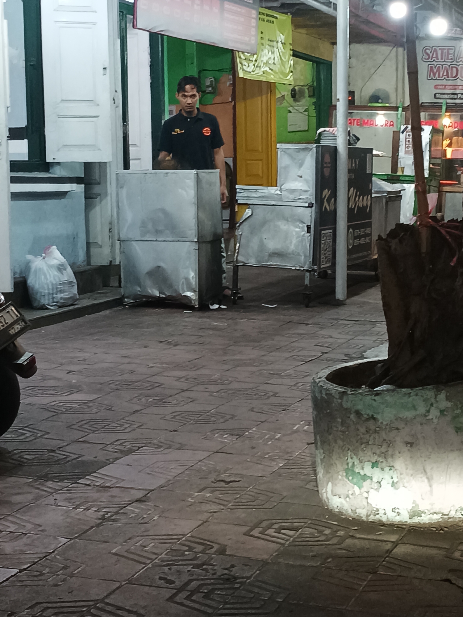 An Indonesian man, wearing a black polo shirt with a logo, stands in front of a stainless steel food stall at night. He appears to be working or attending to customers. The stall is located on a tiled sidewalk in what appears to be a commercial area. In the background, there are signs for "Sate Ayam Madura" (Madura Chicken Satay) and "Ate Malura," suggesting this is a food vendor area. The environment is dimly lit, with overhead lights illuminating the scene. A motorcycle license plate is visible in the foreground, partially obscuring the lower left corner of the image. There is also a large planter to the right, with a tree trunk inside. The overall scene suggests a busy street food environment in Yogyakarta, Indonesia, likely during evening hours.