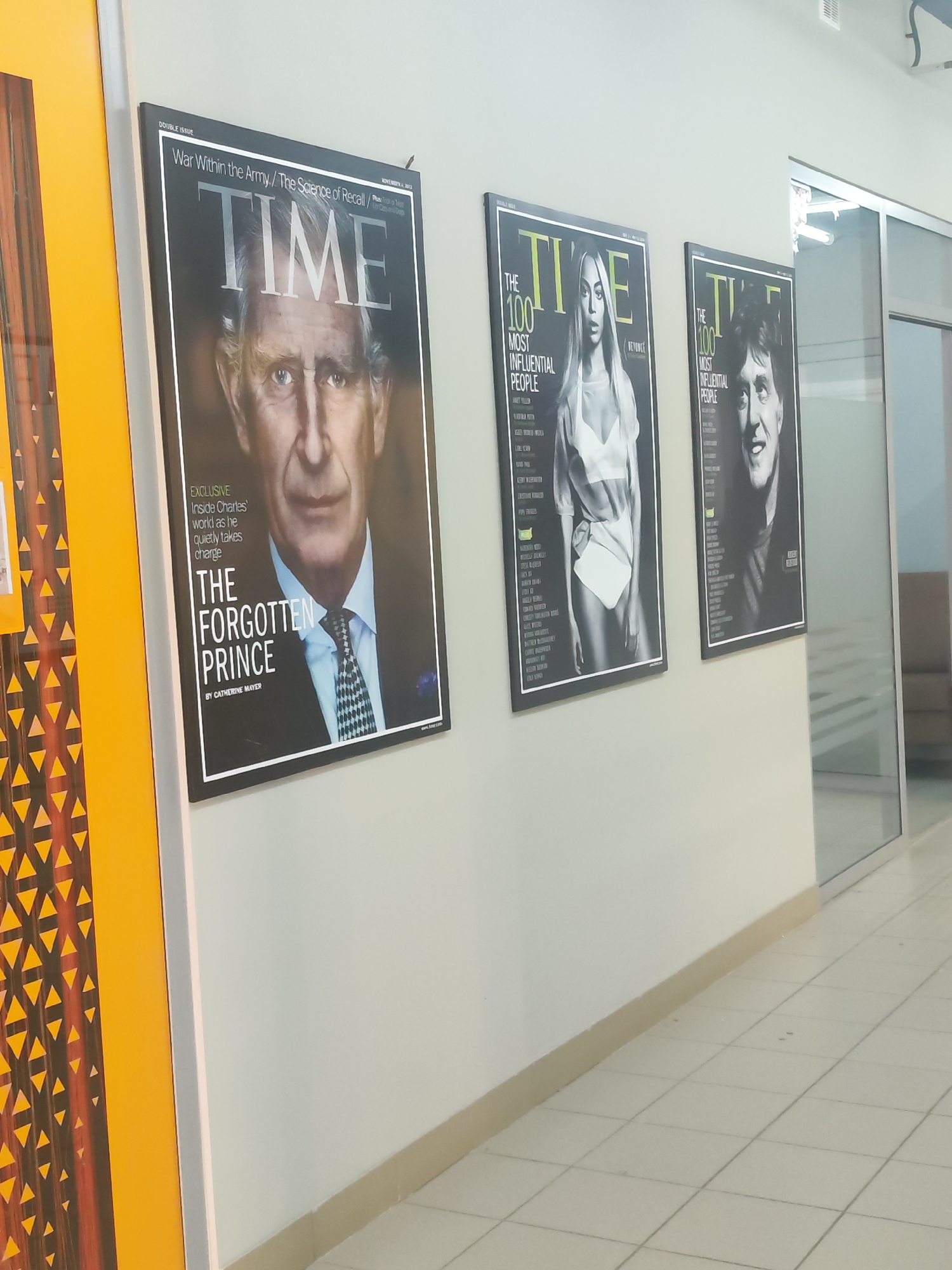 The image shows a hallway with three framed Time magazine covers hanging on a light-colored wall. The cover on the left features Prince Charles with the title "The Forgotten Prince," dating to November 4, 2013. The middle cover shows Beyoncé as one of the 100 most influential people. The cover on the right features an image of Paul McCartney as one of the 100 most influential people. The hallway floor is tiled, and there is a glass-walled room or office to the right of the magazine covers. A yellow door/divider with a geometric pattern is visible on the left side of the image. The scene appears to be an interior hallway, possibly in Chaikovsky, Russia, although there is no visual evidence within the frame to confirm this location.