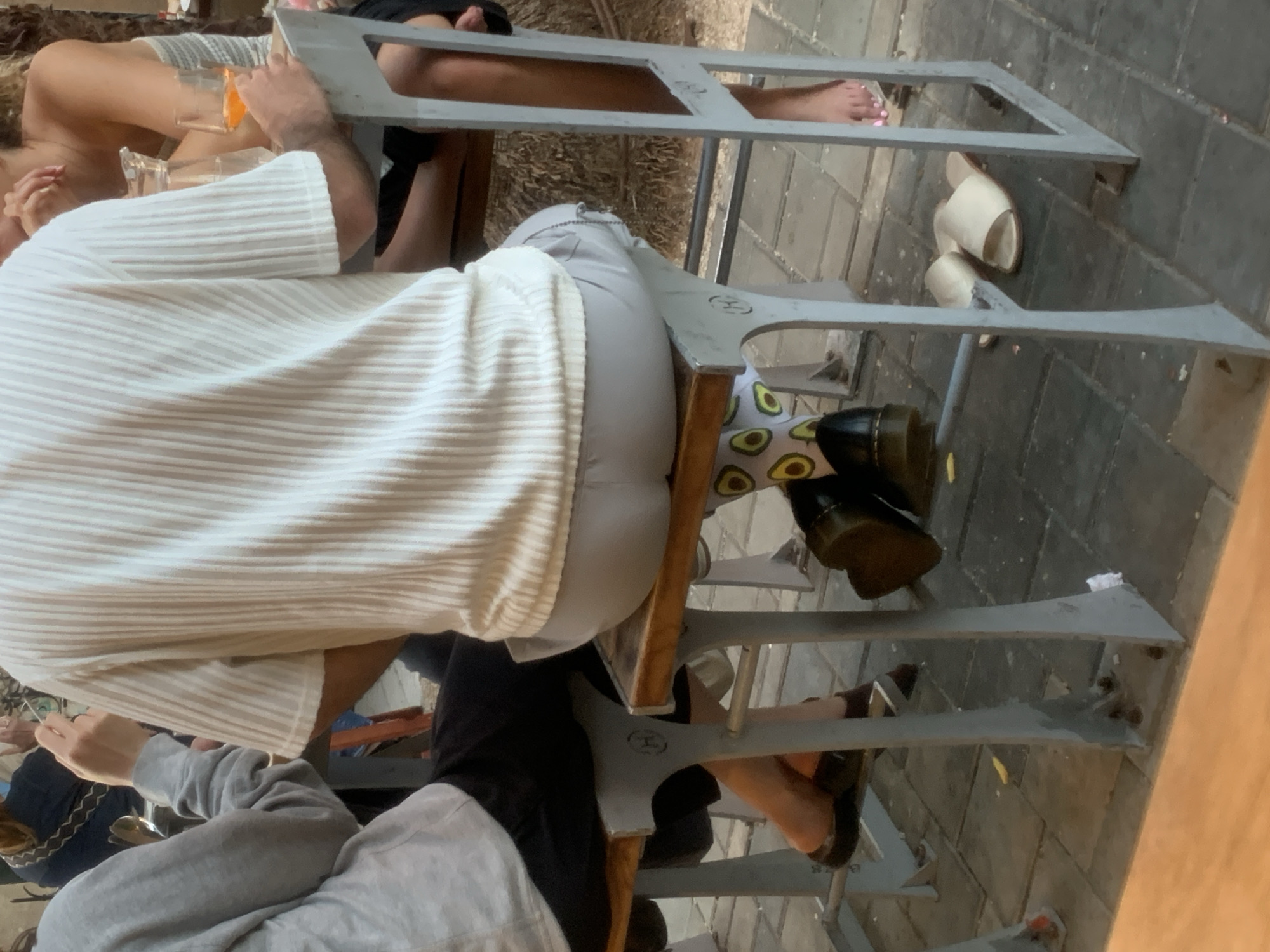 The image shows a group of people seated at tables, seemingly at a restaurant or cafe. One person is wearing a white, horizontally striped shirt and light-colored pants with avocado patterns. This person is seated on a metal chair. The scene appears to be outdoors, evidenced by the brick flooring and the presence of other chairs and tables. The location is likely in Tel Aviv, Israel, based on the visual style of the outdoor space and the presence of details characteristic of the city's street cafes.