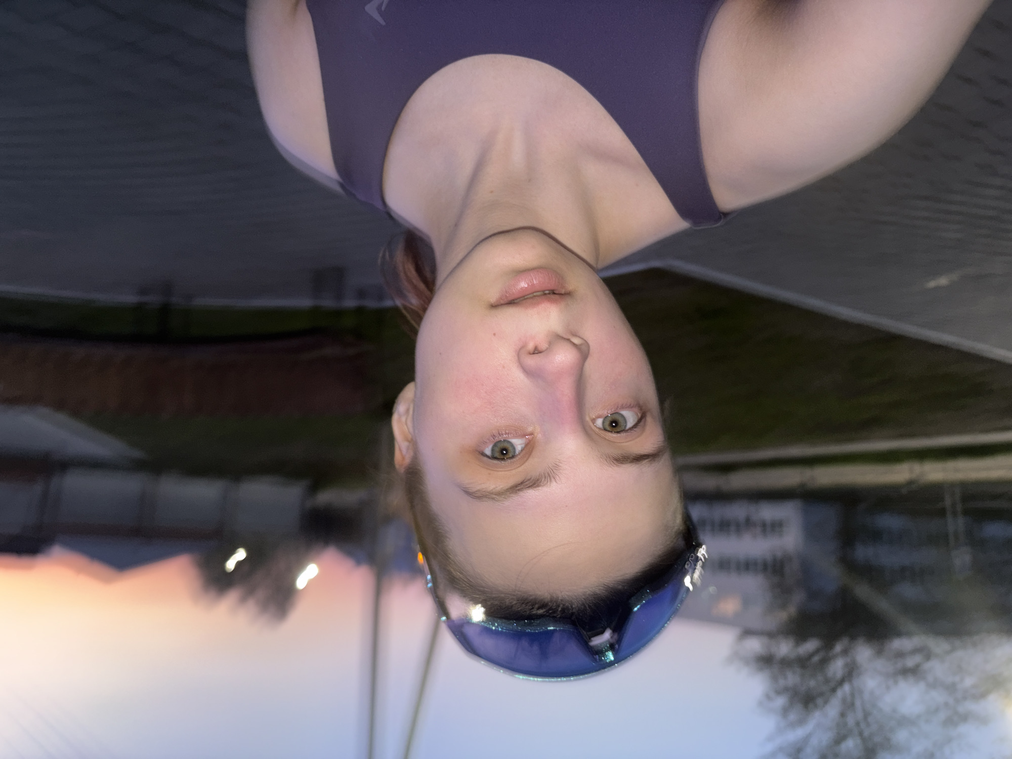 A medium shot depicts a woman from the shoulders up, inverted. She has fair skin, light brown hair pulled back, and light green eyes. She is wearing a dark purple sports bra and blue mirrored sunglasses pushed up onto her forehead. Her expression is neutral.
The background is blurry, appearing to be outdoors at dusk or dawn. There is a pink and orange glow on the horizon, suggesting either sunrise or sunset. Buildings and trees are visible in the background. Based on the prompt, the location is in the city of Прикамский, Russia. The surface beneath the woman's chin looks like a tiled or paved area, possibly a sidewalk or patio.