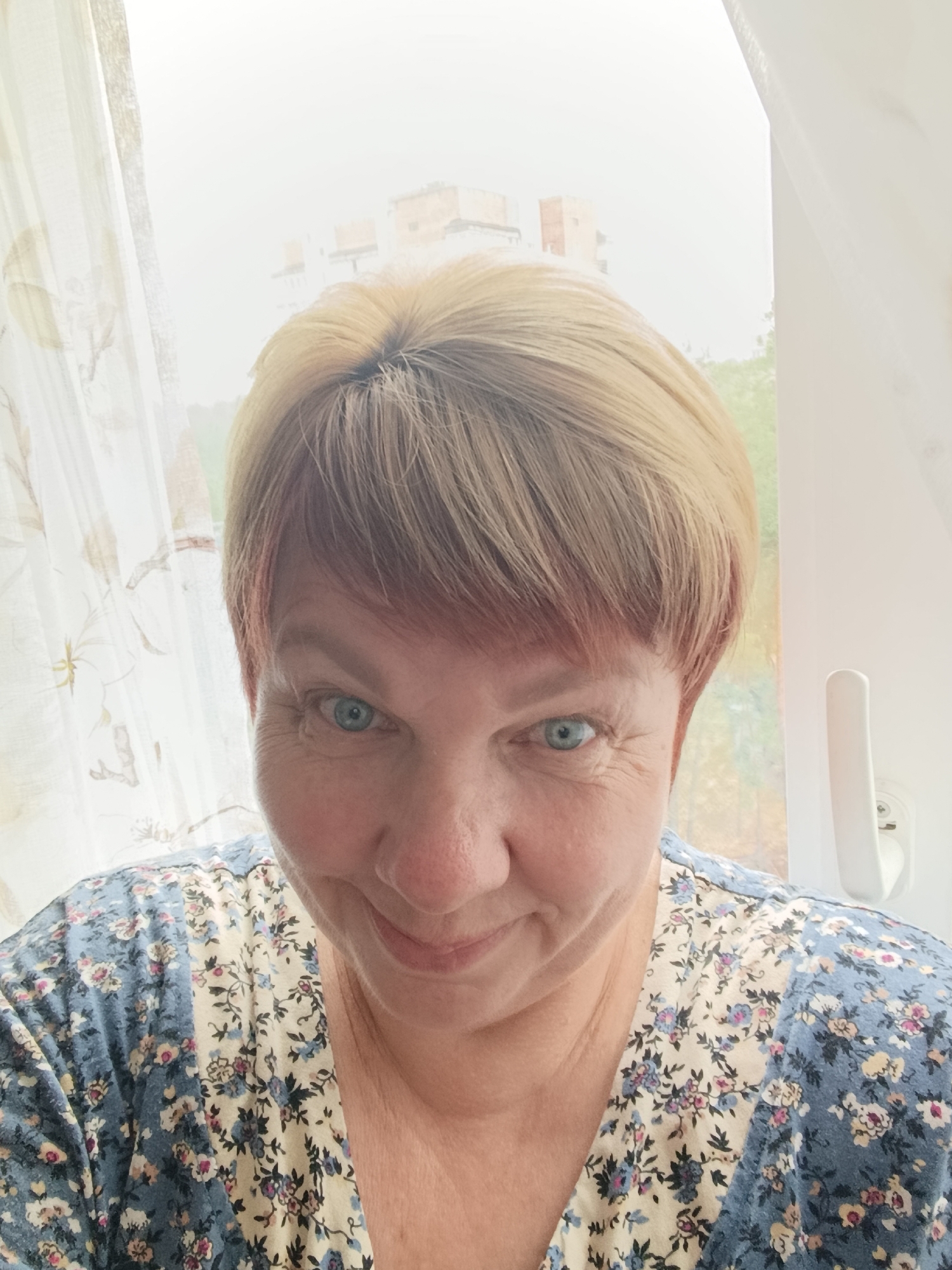 A close-up, selfie-style portrait of a middle-aged woman with short, blonde hair, blue eyes, and fair skin is captured. She is wearing a floral-patterned top with blue and cream colors, featuring small pink and blue flowers. The woman is positioned in front of a window with a sheer, patterned curtain on the left and a white window frame with a handle on the right. The background is bright and somewhat hazy, showing indistinct buildings and trees, suggesting an outdoor environment in an urban setting, potentially Svietlahorsk, Belarus. The woman is looking directly at the camera with a slight smile.