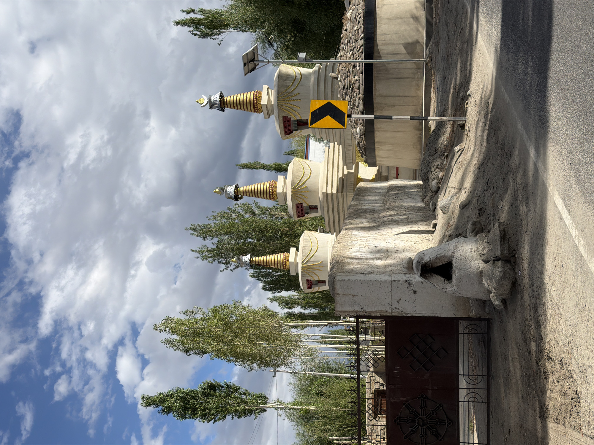 The image depicts a row of three Buddhist stupas with golden finials, situated in the city of Churam, India. The stupas are white and cylindrical, with decorative golden embellishments. They are set against a backdrop of tall, slender trees and a cloudy sky. In the foreground, a dark metal gate with intricate geometric patterns is visible on the left. To the right of the gate, a concrete structure with an opening stands on the ground. A road with a white line marking is present at the bottom of the image. A yellow chevron sign is posted on a striped pole near the stupas. The scene appears to be an outdoor religious site with elements of local architecture and natural surroundings.