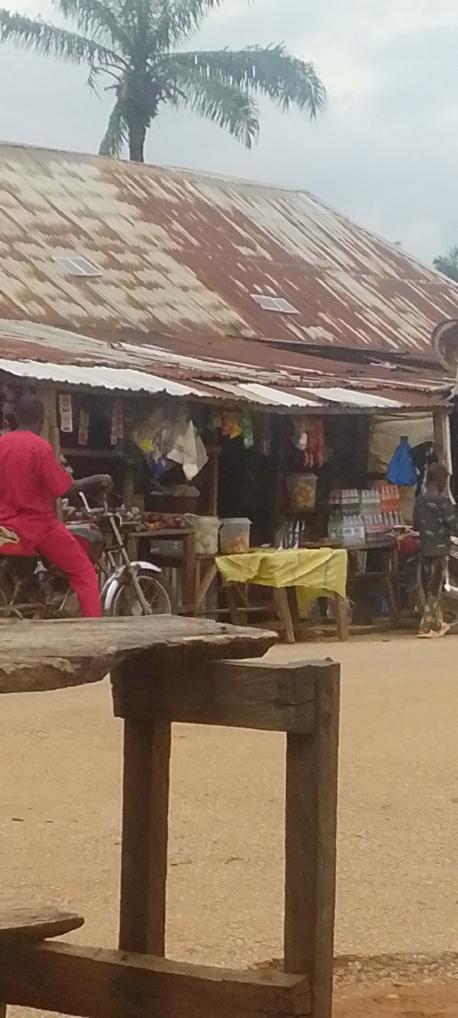 The image depicts a roadside market stall in Baissa, Nigeria. A man wearing a red garment is seated on a motorcycle, facing away from the camera. Behind him, the market stall displays a variety of goods, including bottled drinks and packaged items, on shelves and tables. A yellow cloth is spread over a table in the foreground. To the right of the stall, a person in camouflage clothing stands near another motorcycle. The stall has a corrugated iron roof, with solar panels visible on the upper portion. A palm tree with drooping fronds stands behind and to the left of the stall, with its trunk partially obscured by the roof. The ground appears to be dirt or gravel. The sky is overcast, suggesting a cloudy day. The scene is typical of a local marketplace, with goods laid out for sale and people engaged in daily activities. There is no visible text in the image.