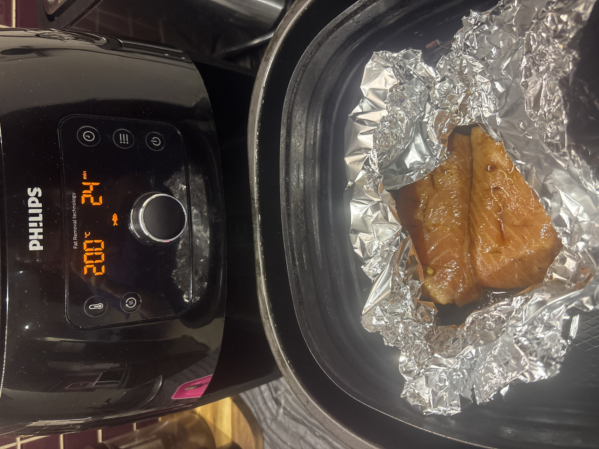The image shows an air fryer set to 200 degrees Celsius for 24 minutes, with two pieces of cooked salmon wrapped in aluminum foil inside the air fryer basket. The air fryer is black with a digital display showing the temperature and time. The salmon appears to be glazed and is glistening. The background shows a kitchen counter and cabinets. There is no information available to determine the location of this image in Dubai, United Arab Emirates.