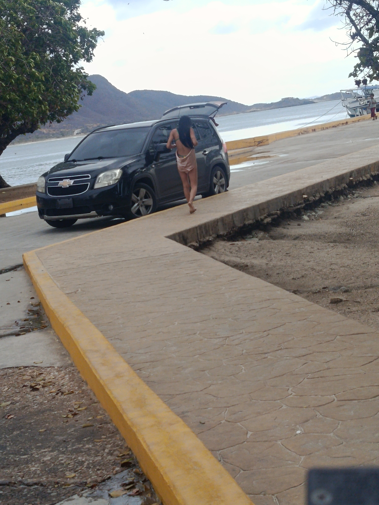 A daytime image captures a scenic coastal or lakeside view in Juan Griego, Venezuela, under a bright, partly cloudy sky. In the foreground, a patterned light-colored concrete path with a raised yellow curb leads into the distance. A dark-colored Chevrolet SUV is parked on a paved road adjacent to the curb on the left, with its rear hatch open. The Chevrolet "bow tie" logo is visible on the vehicle's front grille.
A woman with long dark hair, dressed in a light-colored two-piece outfit, walks barefoot away from the SUV, her back to the viewer, moving towards the right side of the frame. Beyond the road and SUV, a large body of water extends, with mountains rising on the distant shore. Green trees frame the left and right sides of the scene. Several boats are visible on the water, some docked or anchored near the far shore on the right. The terrain immediately to the right of the concrete path is rough and unpaved. The overall scene suggests a leisurely stop or activity in a picturesque waterfront setting.