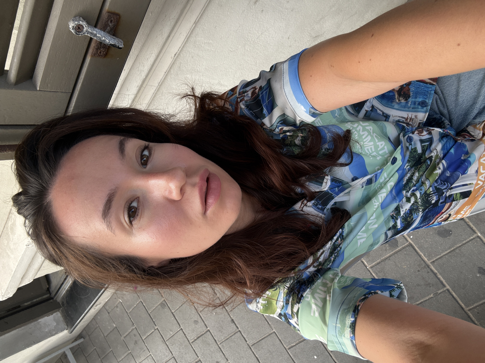 A woman is taking a selfie outdoors. She is wearing a colorful short-sleeved button-down shirt with a print of travel destinations and beaches. Her brown hair is down and frames her face. She is looking directly at the camera with a neutral expression. The background shows a sidewalk made of grey interlocking pavers and a white building with a grey wooden shutter and a metal latch. The scene appears to be in a sunlit urban environment.
