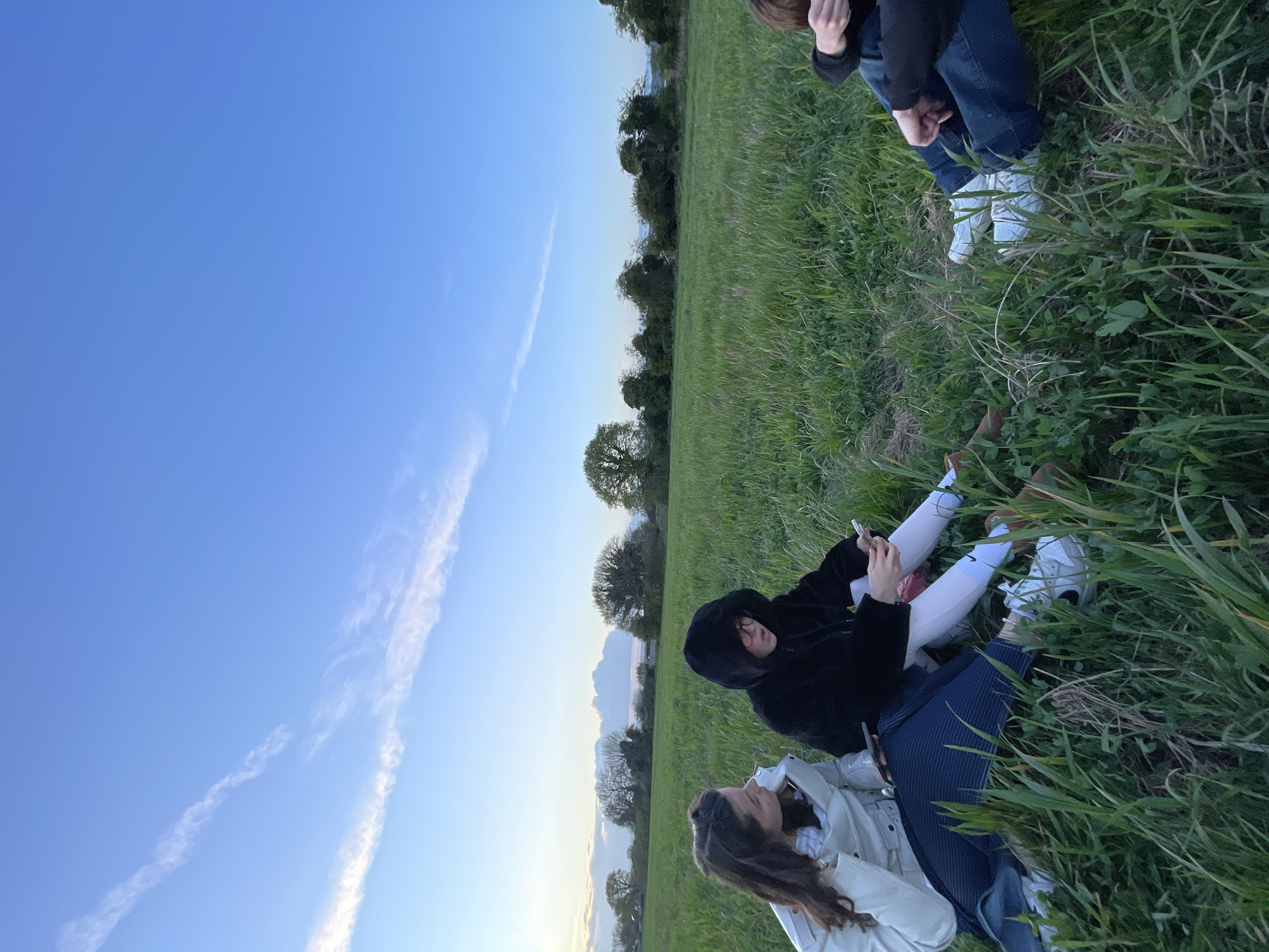 The image depicts a group of people sitting in a grassy field in Maidstone, United Kingdom. Two individuals are positioned in the lower center of the frame, seemingly relaxing on a blanket. One is wearing a light-colored jacket, and the other is wearing a dark hooded jacket and white pants, possibly using a phone. To the right and slightly above them, there are two more people, only their legs and partial torsos are visible. These individuals wear jeans and white shoes. The background includes a clear blue sky with contrails, a tree line, and green fields. The photograph seems to be taken during late afternoon or early evening, judging by the sky's hue. The image appears rotated.