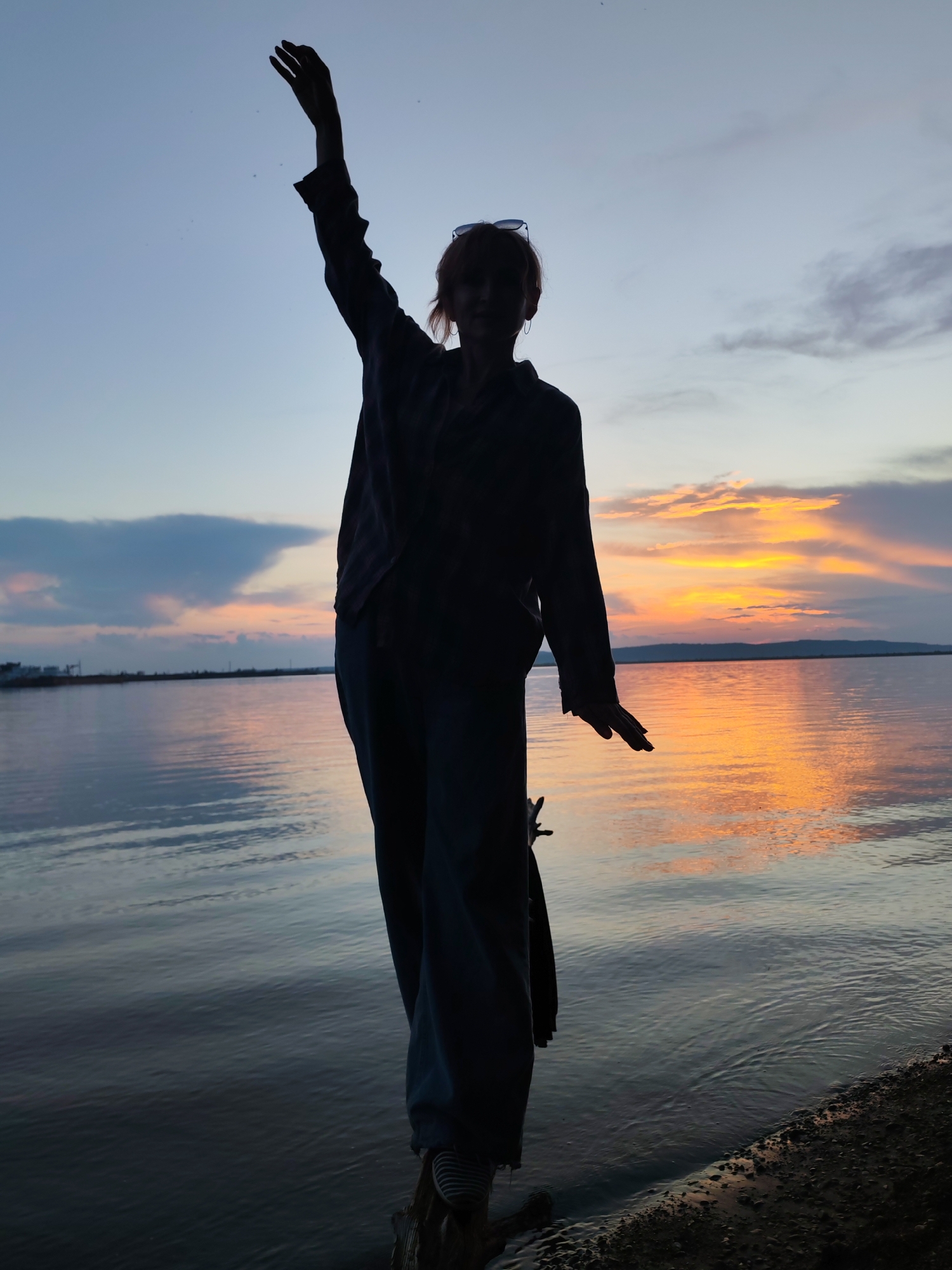 A full-body, silhouetted shot captures a person standing on a wooden structure near the water's edge at sunset. The individual is facing the camera, with their right arm raised and bent at the elbow, palm facing forward. Their left arm is extended to their side, bent at the elbow, with their hand facing downwards. The person is wearing a long-sleeved collared shirt and loose-fitting pants. Their hair is short, and sunglasses are perched on their head. The background is dominated by a body of water, reflecting the vibrant colors of the sunset. The sky displays a gradient of blue, orange, and yellow hues, with scattered clouds. In the distance, a faint outline of a city or industrial area is visible across the water. The scene is set during the golden hour, emphasizing the silhouette against the dramatic sky and the calm water. This location is likely the Kama River or a reservoir in the city of Chaikovsky, Russia, known for its picturesque sunsets over the water.