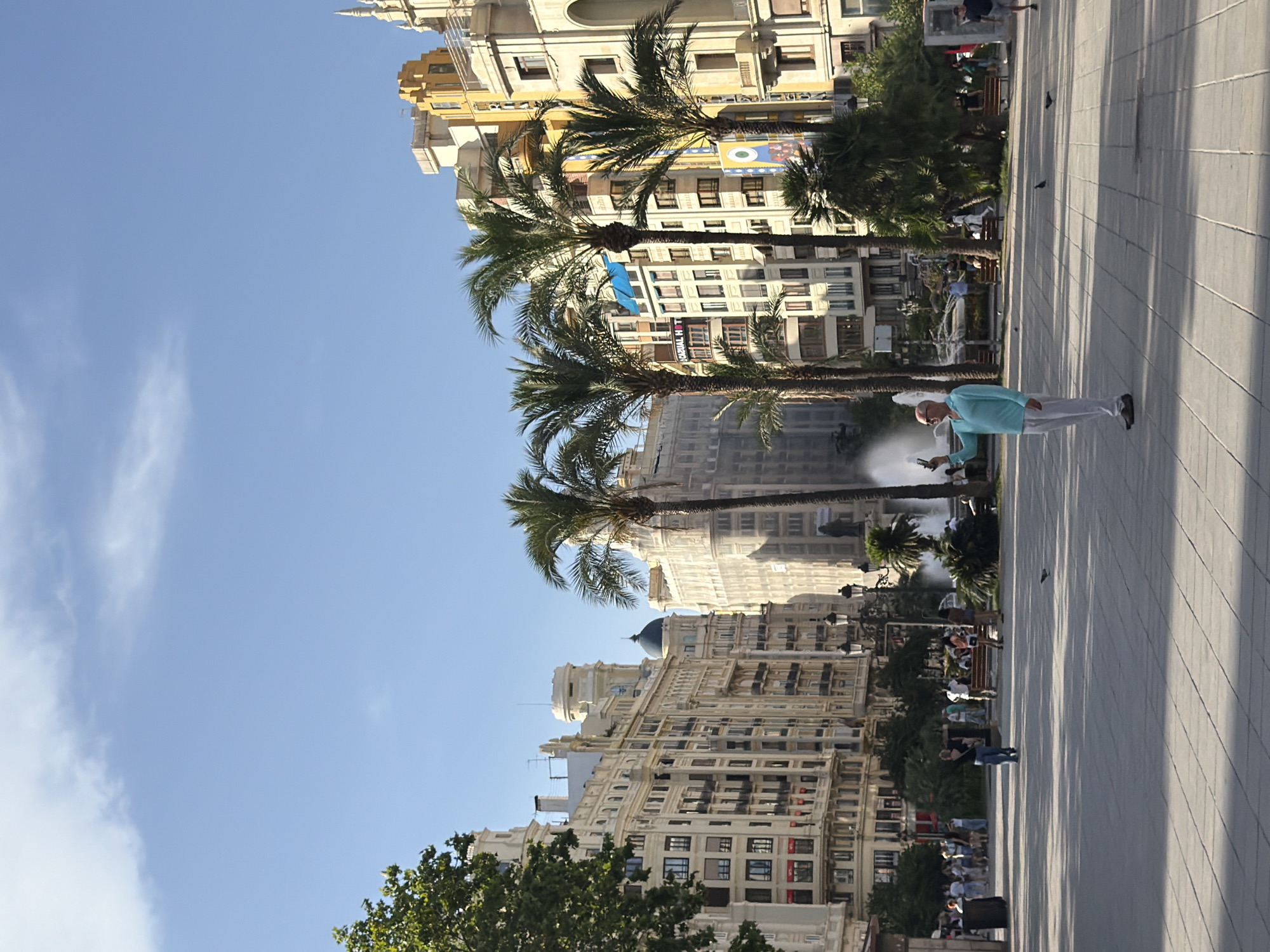 An eye-level, full outdoor shot captures a sunny day in a European city square. A bald, white man in a light blue long-sleeved shirt and white pants stands in the foreground, holding a smartphone horizontally as if taking a picture of himself or the surroundings. Palm trees are scattered throughout the scene, with several tall ones framing the view of ornate, pale buildings in the background. The architecture suggests a classic European style, with detailed facades and multiple stories. A fountain is visible in the mid-ground, with water spraying upwards. Several pigeons are on the tiled ground, and a few people are scattered throughout the square, some sitting on benches and others walking. The sky is a clear blue with a few wispy clouds. The sun casts long shadows across the square, indicating a late afternoon setting.