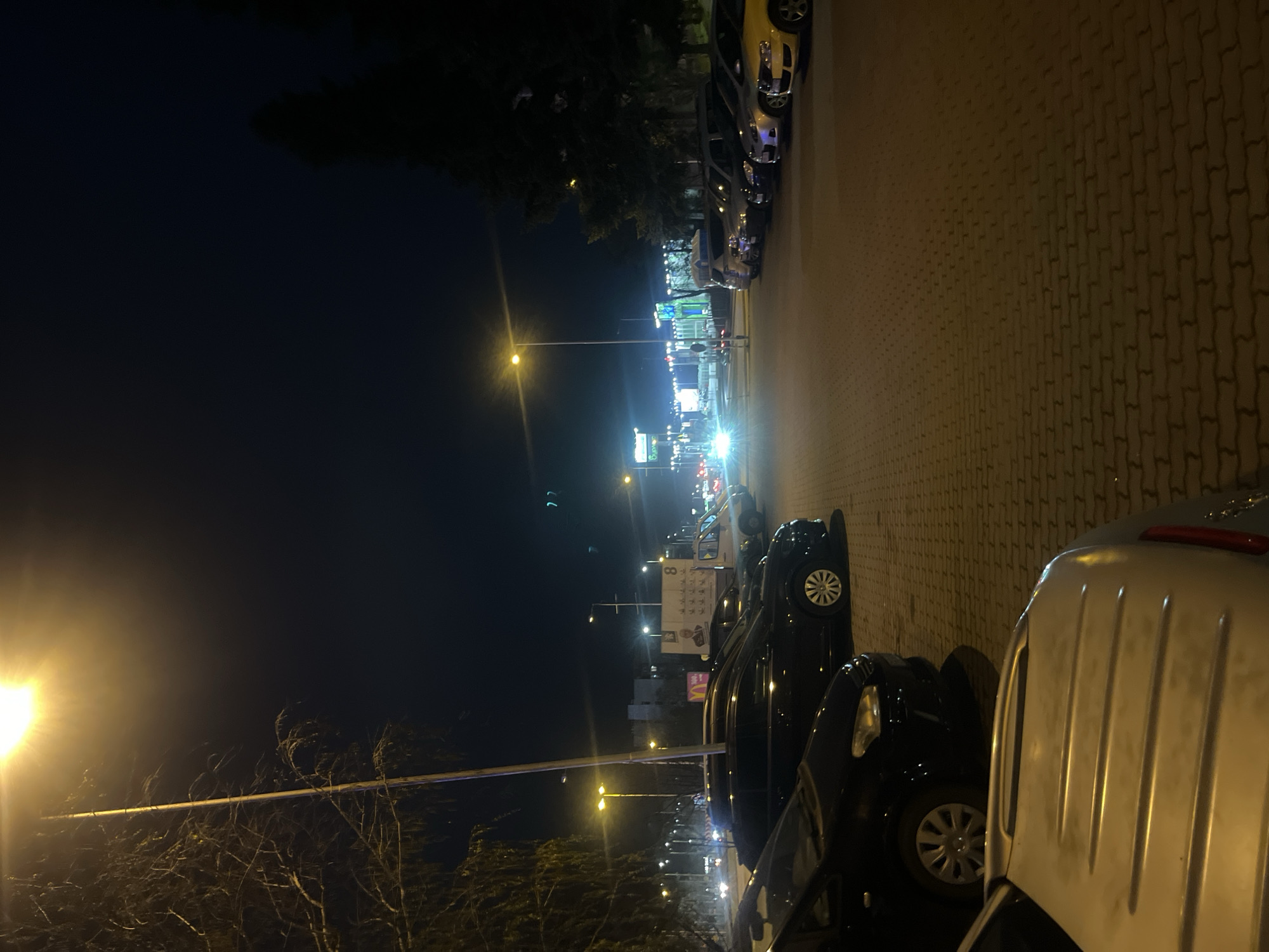 The image depicts a street in Veliko Tarnovo, Bulgaria at night. The view is from a low angle, oriented vertically. The road is paved with interlocking blocks. Cars are parked along the right side of the street, facing away from the viewer. Streetlights illuminate the scene, with more intense light coming from further down the street where there are shops and businesses. In the distance, a sign with "8" and other illuminated advertisements are visible. The sky is dark, and trees are visible along the left side of the street, silhouetted against the night sky and streetlights. No people are clearly visible. The dominant color in the image is yellow-orange from the street lights.