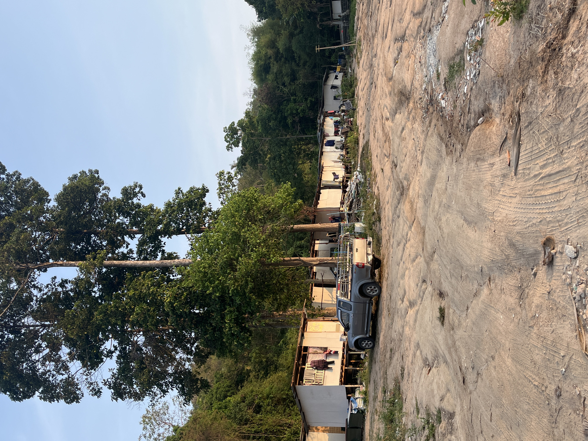 The image captures a rural or semi-rural scene in Baan Nok Sai, Thailand, featuring a dirt road alongside simple dwellings, backed by lush green hills.
In the foreground and midground, a wide, unpaved dirt path extends, marked extensively with tire tracks and some footprints. Scattered debris, including broken white shards (possibly glass or ceramic) and natural organic matter, is visible across the ground. A gray pickup truck with a metal utility rack in its bed is parked on the dirt path on the right side of the frame.
To the left of the path, a series of small, single-story structures, resembling simple shacks or temporary housing units, are lined up. These dwellings appear to be constructed from light-colored materials. Several items of laundry are seen hanging outside the buildings, suggesting active occupancy.
Large, mature trees with prominent trunks and dense green canopies line the left side of the scene, reaching high towards the sky. Behind the structures, a steep hillside covered in dense, dark green tropical foliage rises, forming a natural, verdant backdrop that stretches into the distance.
The sky above is clear and light blue, indicating fair weather. The quality of light suggests it is daytime, possibly late afternoon. No people are visible, and there is no discernible text in the image. The overall impression is one of a functional, lived-in area situated within a natural, heavily vegetated environment.