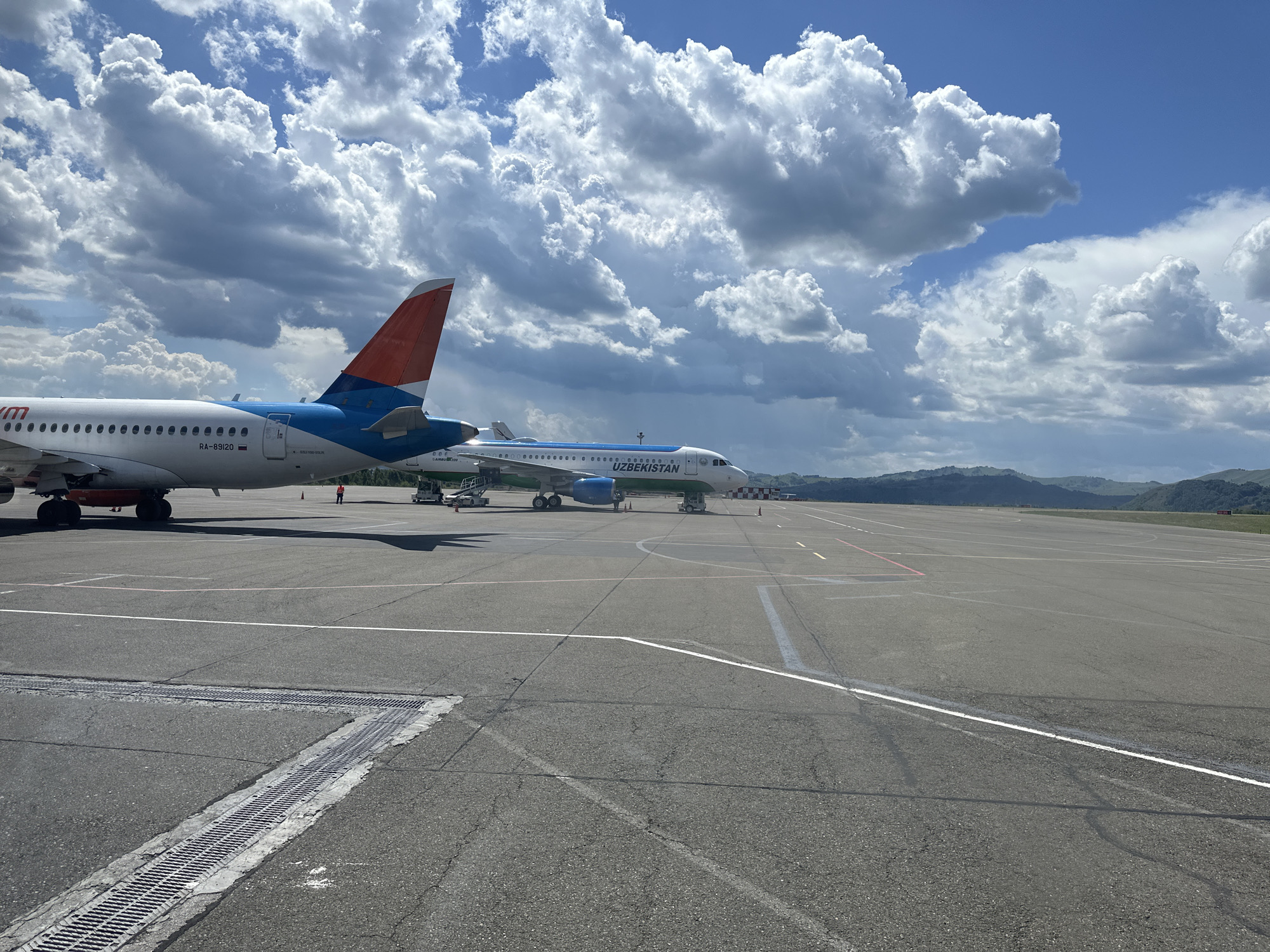 The image depicts an outdoor scene at an airport, likely in Maima, Russia. Two airplanes are parked on the tarmac. The airplane on the left has a red and blue tail with the registration "RA-89120" visible. The airplane on the right is white with green and blue accents and the word "UZBEKISTAN" written on its side. The tarmac is paved and marked with white and red lines. In the background, there are hills and mountains under a partly cloudy sky. The image is captured from a low angle, showing a portion of the tarmac in the foreground with drainage grates. There are no visible actions or interactions occurring between people or objects.