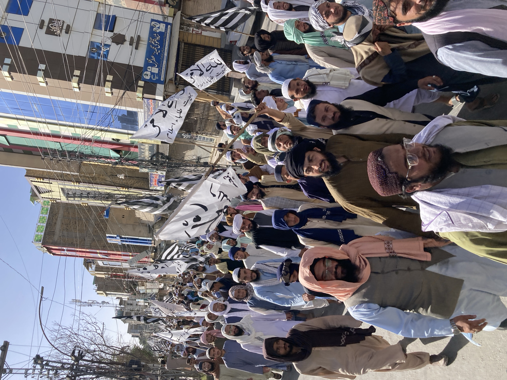 A large crowd of people, predominantly men with beards and wearing traditional Islamic clothing and headwear, are marching down a street in Quetta City Tehsil, Pakistan. Many of them are holding white flags with Arabic writing and black and white striped flags. The street is lined with buildings, including a building that has a sign indicating that it's a "QARAQULI CAP HOUSE". Power lines and poles are visible above the crowd. The atmosphere appears to be a demonstration or rally. The photograph is taken during daytime with clear skies.