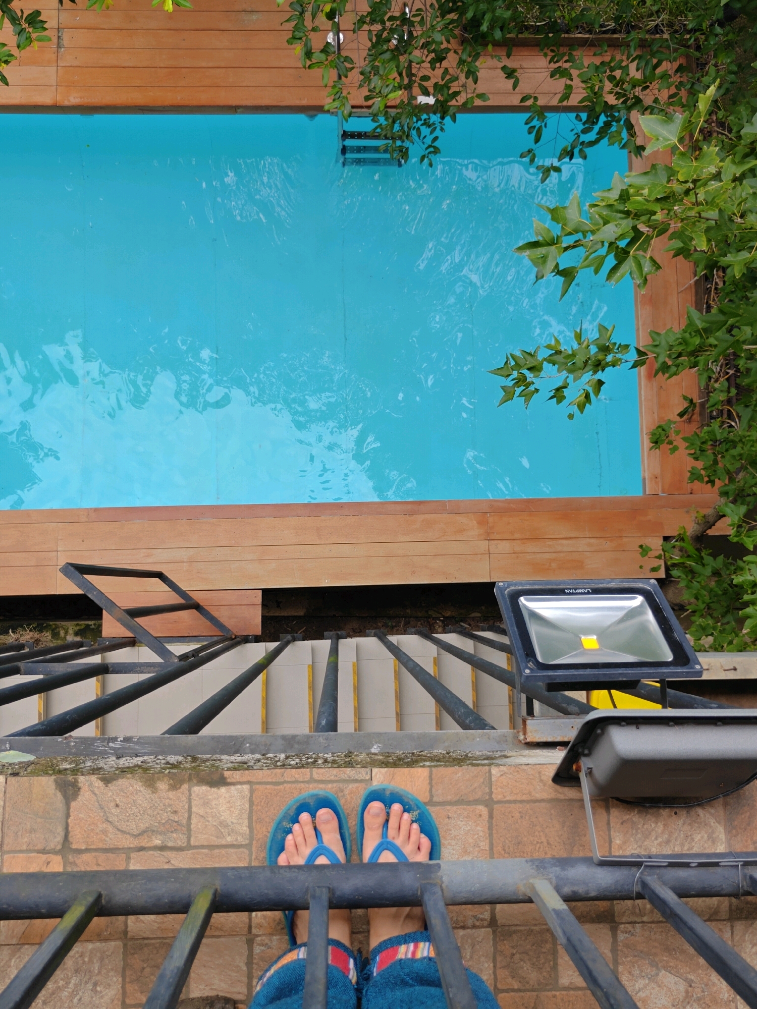 An overhead, eye-level shot shows a person's feet wearing blue flip-flops and blue fuzzy slippers, standing on a tiled patio. In front of the patio is a dark metal railing. Behind the patio, a set of stairs with yellow markings on the edge of each step descends, partially obscured by the metal railing. Further back is a swimming pool with bright blue water, surrounded by a wooden deck. Green leaves from trees hang over the pool and deck. A floodlight is mounted on a pole to the right of the stairs. The scene appears to be an outdoor residential area, possibly a private home with a swimming pool, located in Mu Si, Thailand.
