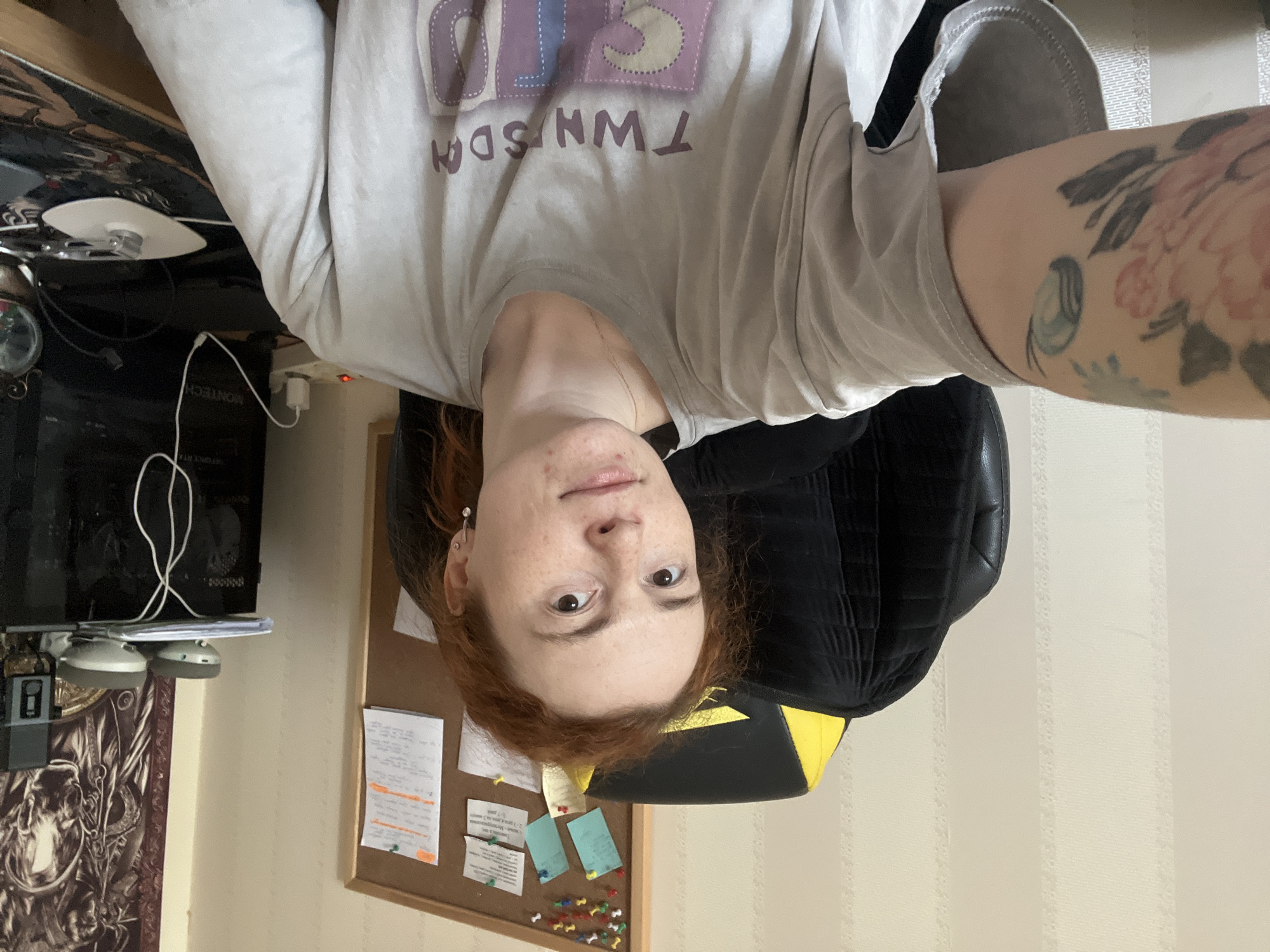 A rotated selfie-style image shows a red-haired woman with freckles and brown eyes, wearing a light gray t-shirt with the word "TWNSDA" printed on it. The woman is seated in a black chair with yellow accents. She has a tattoo on her right forearm. Behind her, there is a cork bulletin board with several notes and papers pinned to it. To her left, part of a computer tower and accessories are visible on a desk. The location is implied to be in Chaikovsky, Russia.