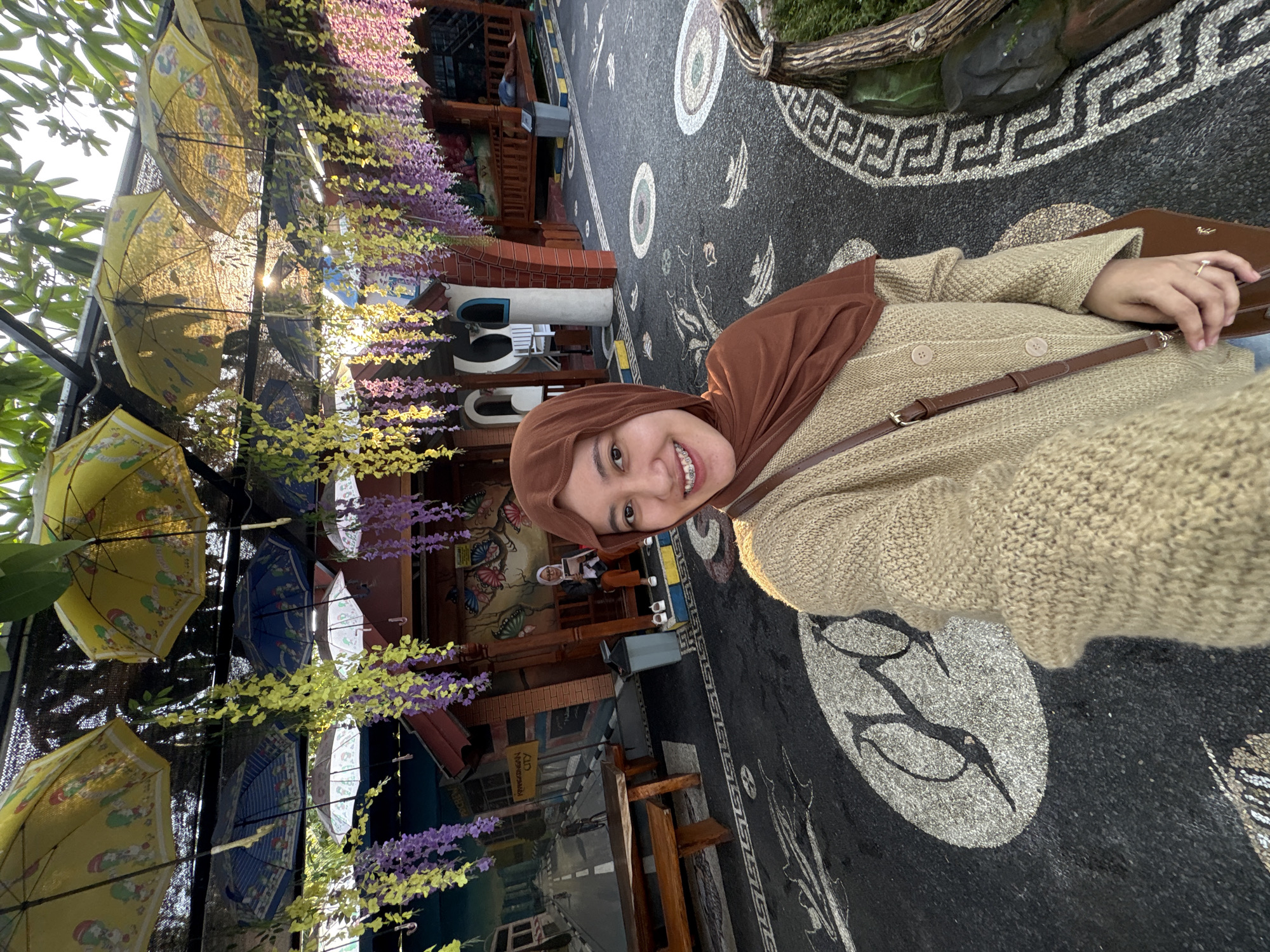 A young woman wearing a brown hijab and a light brown knitted cardigan is taking a selfie outdoors. She is smiling and her braces are visible. A brown crossbody bag is slung over her shoulder. The background appears to be an outdoor seating area decorated with numerous colorful umbrellas hanging overhead and artificial floral garlands. The ground is paved with dark material and features decorative stone mosaic patterns, including what looks like stylized glasses and fish. In the background, there's a mural on a wall depicting a street scene, some brick structures, and outdoor furniture. A second woman, also wearing a hijab, is visible sitting on a bench in the background, reading a book. The scene is set in Kebonsari, Indonesia.