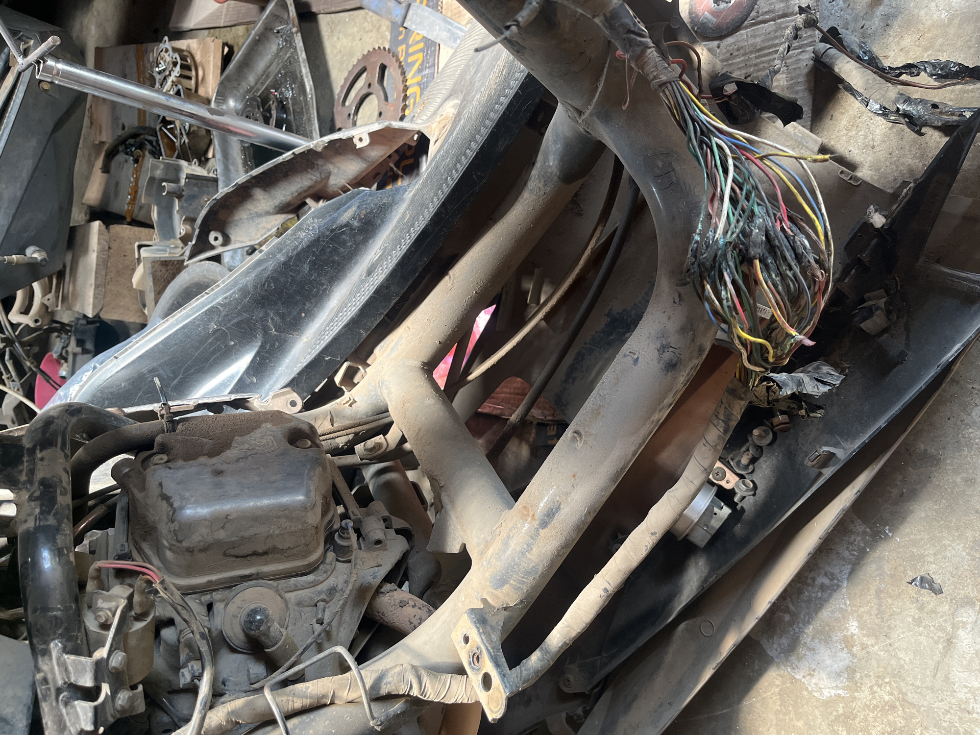 This is a close-up, eye-level photograph of a partially disassembled motorcycle engine and its wiring harness, likely in a workshop or storage area in Kebumen, Indonesia. The scene is cluttered and dusty, indicating neglect or ongoing repair work. The motorcycle components are primarily made of metal and plastic, with various wires and cables visible, many of which appear damaged or exposed. The lighting is natural, coming from an unseen source, casting shadows and highlighting the grime on the surfaces. No individuals are present in the frame.