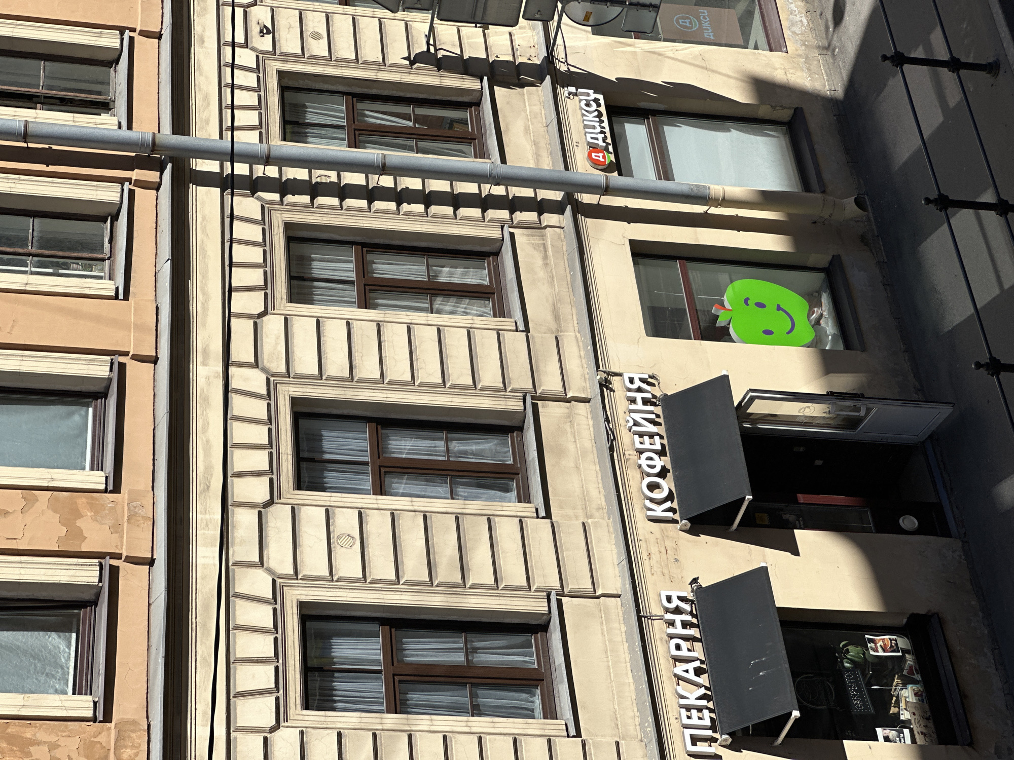 The image presents an elevated, angled view of a multi-story building facade during daytime, illuminated by strong sunlight from the left.
The building's upper section features two visible levels. The topmost level exhibits an older, textured facade with peeling, light orange paint. Two windows are partially visible, set within simple frames. Below this, a horizontal gray ledge separates it from a second level with a lighter, beige-colored facade. This second level displays more ornate architecture, including three windows with dark frames and light-colored sheer curtains, each set within decorative rectangular surrounds. Vertical, fluted pilasters separate these windows. A thin, dark horizontal line or wire runs across this section.
The lower section of the building, at street level, continues the light beige facade. It houses three commercial establishments. From left to right:
1. **ПЕКАРНЯ (Bakery):** Features white, raised Cyrillic lettering "ПЕКАРНЯ" above a black retractable awning. Below the awning is a large display window showcasing various items, including a green plant and what appears to be promotional material, with the word "ОТКРЫТО" (OPEN) faintly visible.
2. **КОФЕЙНЯ (Coffee Shop):** Identified by white, raised Cyrillic lettering "КОФЕЙНЯ" above a black retractable awning. Its glass-paneled door is open, revealing a dark interior.
3. **ДИКСИ (Dixy Supermarket):** Marked by a red and white sign with Cyrillic lettering "ДИКСИ" and a distinct red 'Д' logo incorporating a green circle. This establishment has large glass display windows. The leftmost window prominently features a large, bright green cutout of a smiling apple.
A gray metallic downspout runs vertically down the building's facade, starting from the upper levels and extending to the street level, positioned between the coffee shop and the Dixy supermarket.
The street-level pavement appears dark grey. Strong, well-defined shadows are cast across the pavement by the awnings, the downspout, and an iron railing with vertical balusters visible in the bottom right corner of the frame. Portions of street signs or a utility pole are partially visible in the upper right.
The presence of Cyrillic text "ПЕКАРНЯ," "КОФЕЙНЯ," and the recognizable "ДИКСИ" brand indicates that the location is in a Russian-speaking country, most likely Russia. The specific city and region are unknown.