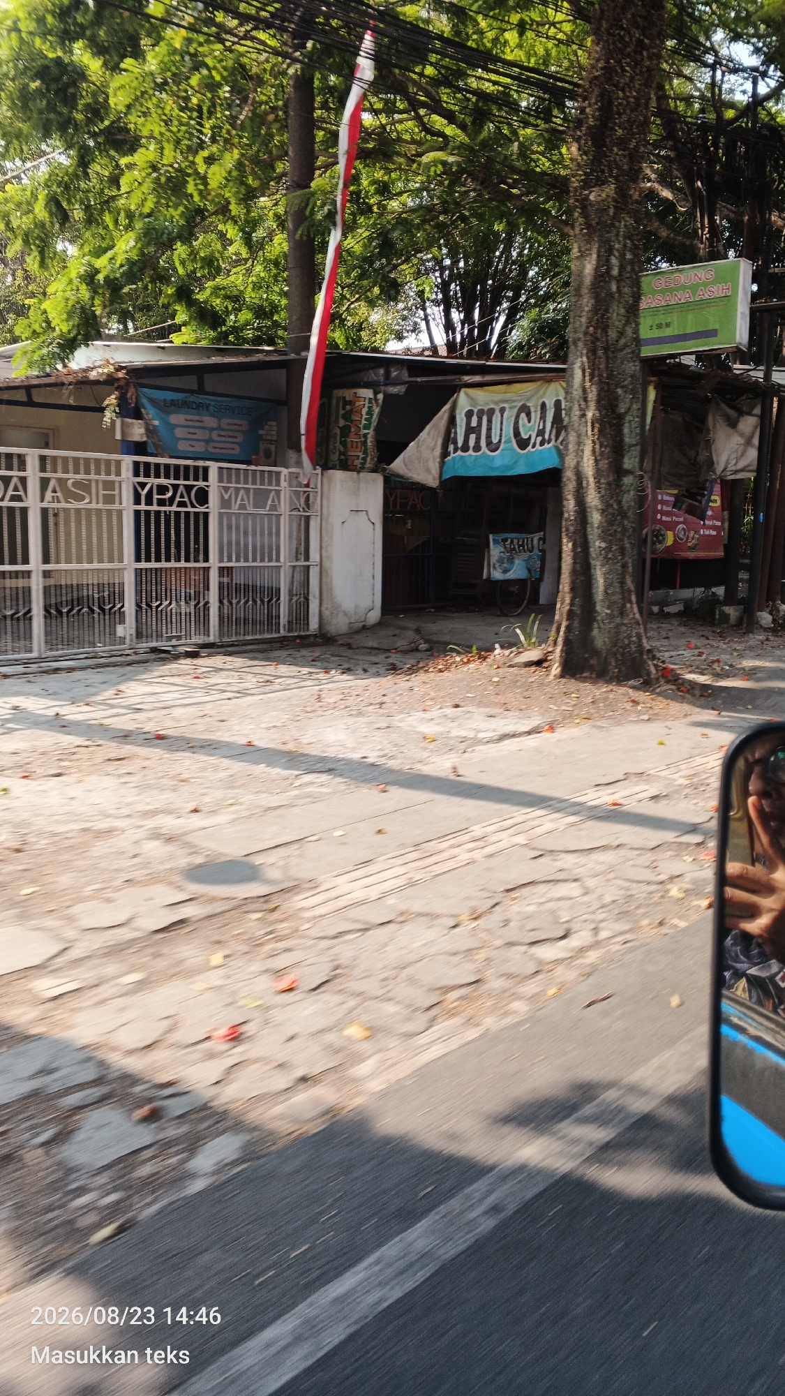 The image captures a street view in Bunulrejo, Indonesia. A building with a white metal gate is visible, with the text "DA ASIH YPAC MALANG" partially obscured by the gate. To the right, a small shop with a blue awning displaying "TAHU CAM" is present, along with a white cart. A green sign with "GEDUNG SASANA ASIH" is also visible in the background. A red and white Indonesian flag is hanging from a pole. The ground is paved with concrete and asphalt, with some fallen leaves scattered about. A large tree stands in the foreground. In the lower right corner, a reflection of a person's hand and arm is visible in a car's side mirror. The timestamp "2026/08/23 14:46" and the text "Masukkan teks" are overlaid on the image.