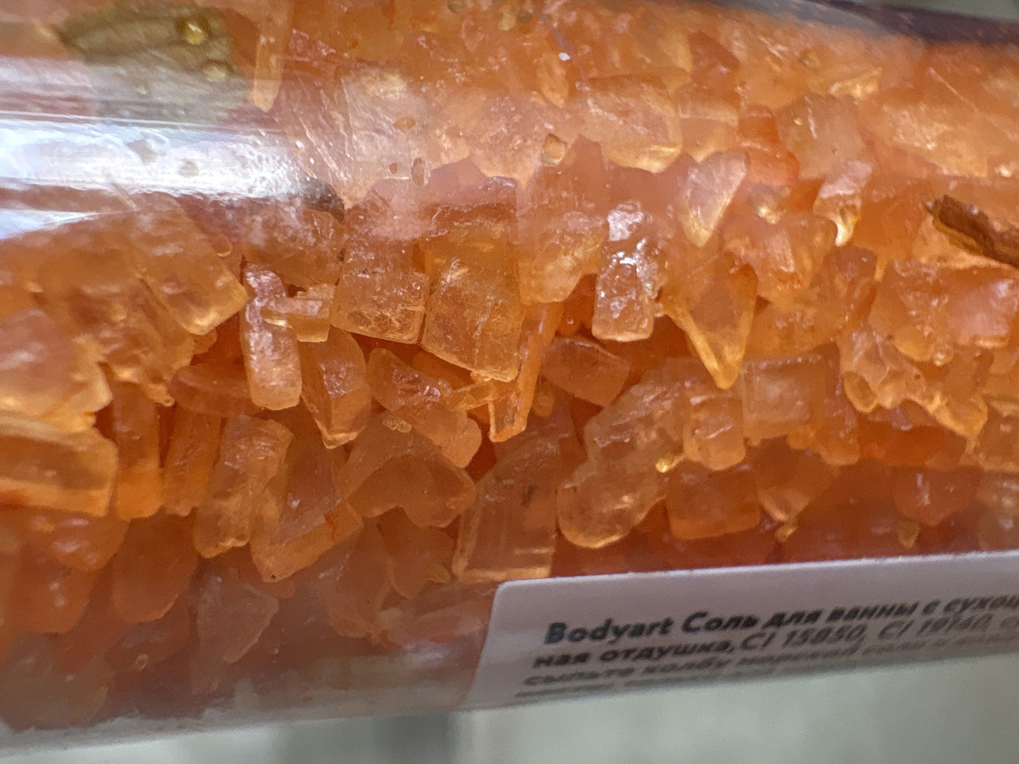 A close-up shot of a container filled with orange, crystal-like bath salts or granules. A label is partially visible at the bottom, with text in Russian: "Bodyart Соль для ванны с сухоц ная отдушка, СІ 15850, СІ 19140". The context suggests that the location is Chaikovsky, Russia.