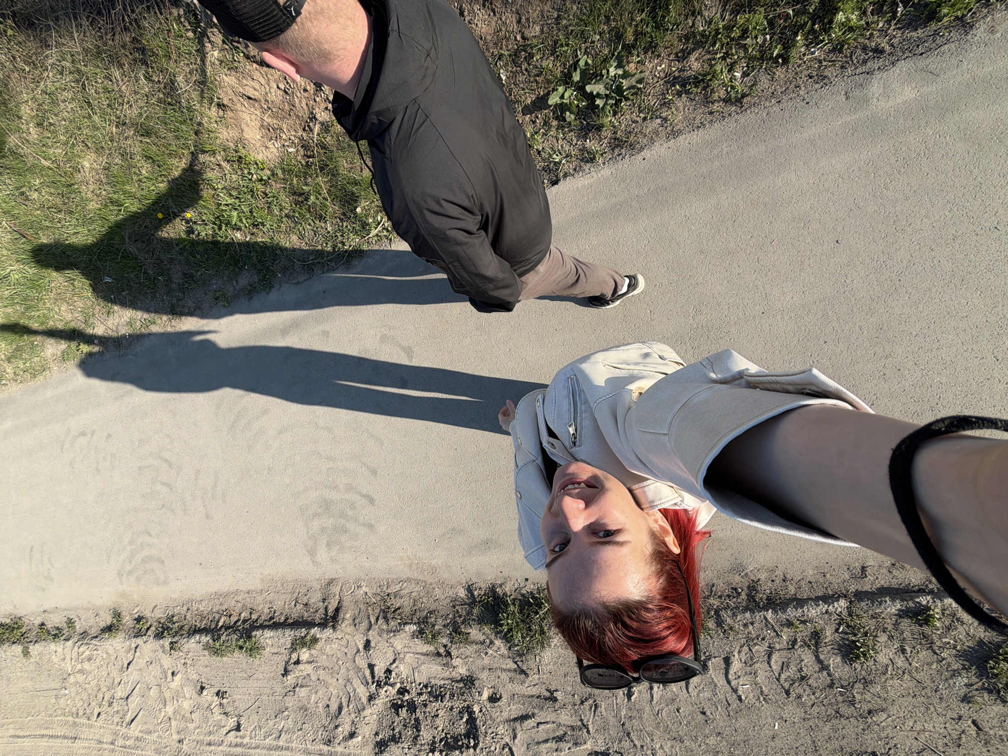 A high-angle selfie captures a red-haired woman with sunglasses perched on her head. She is wearing a light-colored jacket. Beside her and slightly above is a man with short blond hair, wearing a dark hooded jacket and beige pants with dark sneakers. They stand on an asphalt path bordering grassy and sandy ground. Shadows of the people are visible on the path. The location is identified as the city of Chaikovsky, Russia, based on geographical contextual clues.
