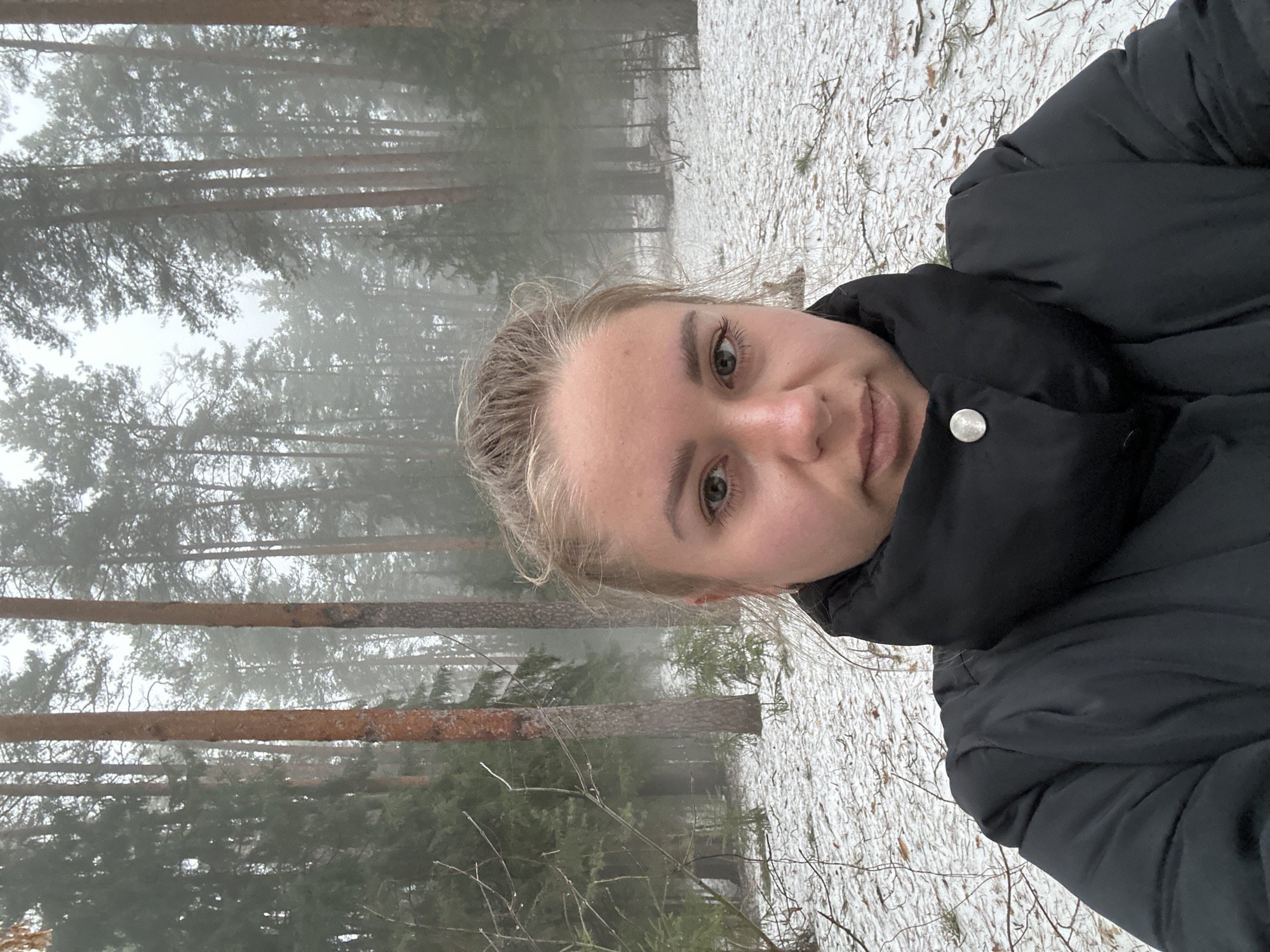 A person with fair skin and blonde hair stands in a foggy forest landscape. Snow lightly covers the ground. The person wears a black puffer jacket. Tall pine trees form the backdrop, fading into the fog. The location is likely in a forest near Chaikovsky, Russia, based on the likely location of the photo's origin.