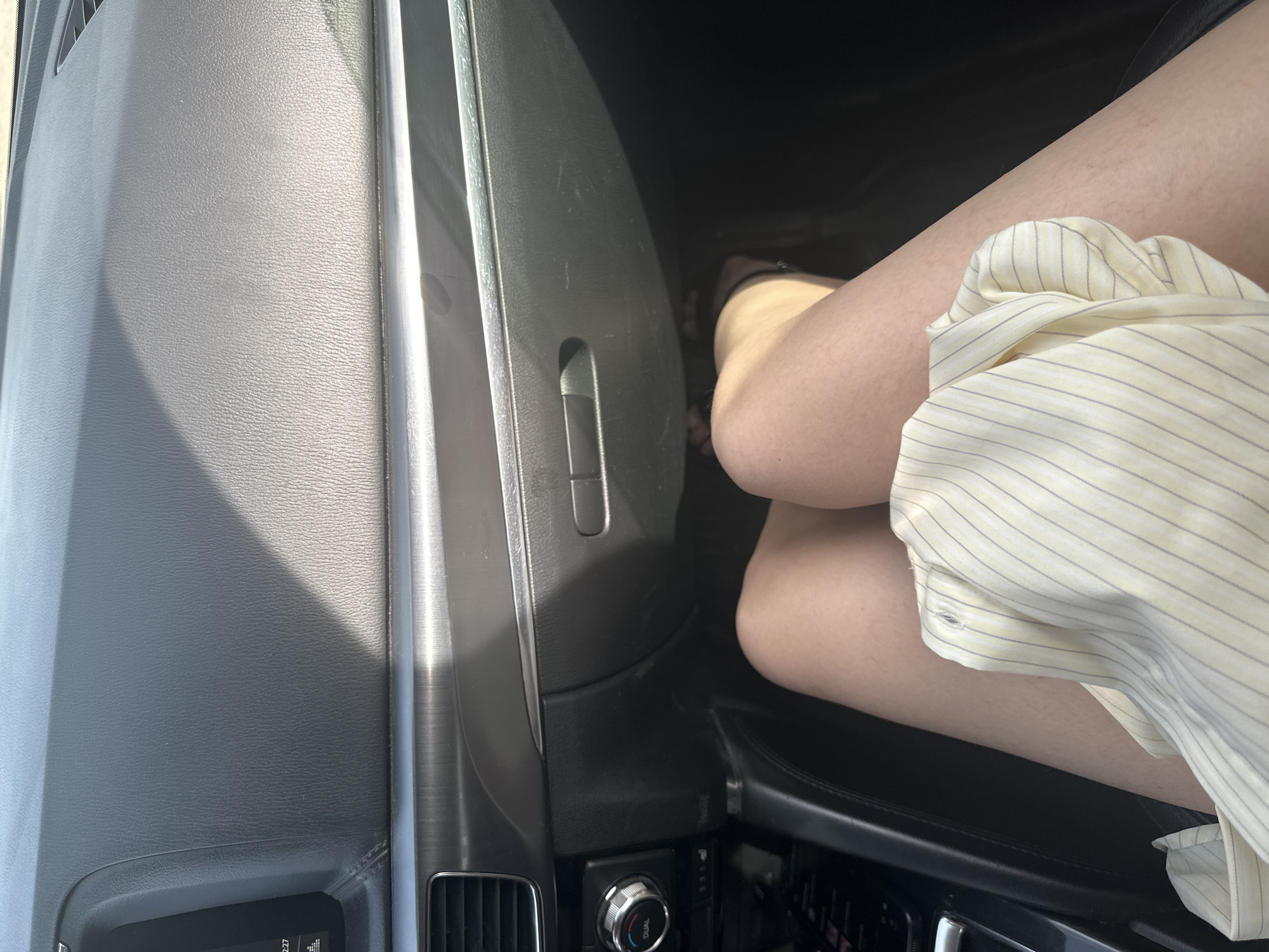 The image shows a portion of a car's interior, focusing on the dashboard and passenger area. The dashboard is dark gray, and the passenger's legs are visible. The passenger is wearing a light yellow striped shirt. The car's climate control panel and some interior trim are visible. The location is unknown.