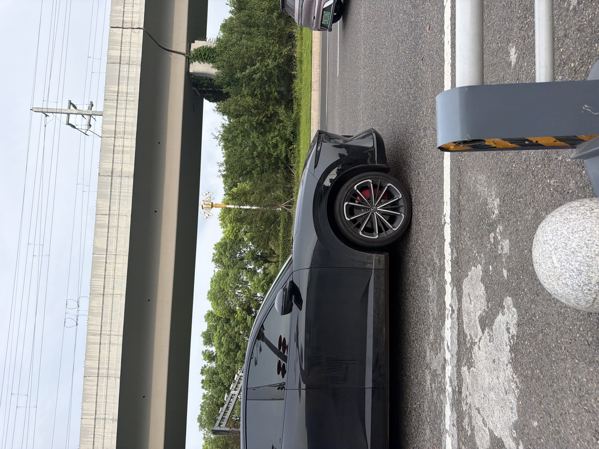 The image depicts a street scene in the city of Jiangjin, China. In the foreground, a black car is parked, partially obscuring a grey guardrail with yellow and black hazard markings. The road surface shows signs of wear, with white lane markings and patches of discolored asphalt. In the midground, a light-colored car with a license plate reading "A 0R8178" is driving away. Behind the cars, a lush green expanse of trees lines the roadside, leading up to a concrete overpass. The overpass is illuminated by streetlights, and power lines are visible above. The sky is overcast, with a few wispy clouds. The overall impression is of a typical urban landscape, with elements of infrastructure and nature coexisting.