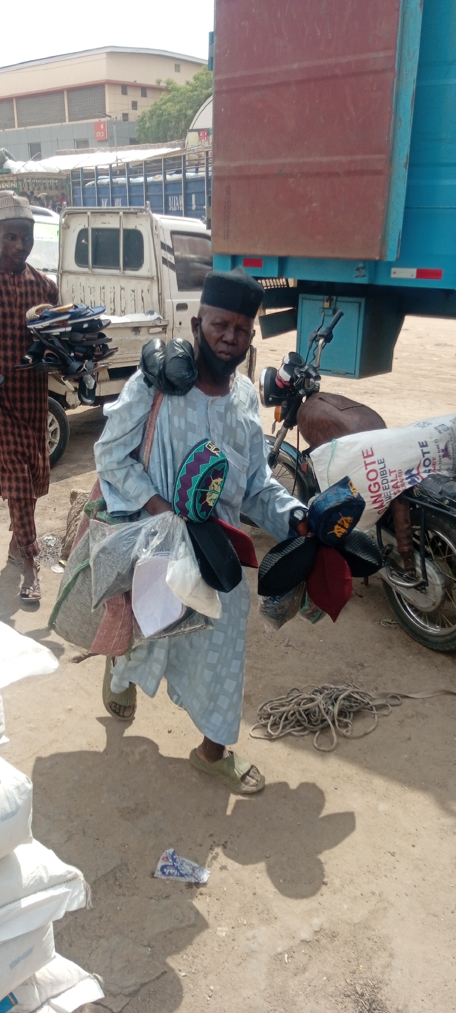 The image shows a man in Sabon Gari West, Nigeria, carrying various items for sale. He wears a black cap and a light blue patterned robe. He has bags and items slung over his shoulder and held in his hands, including hats, sandals, and other bags. A man wearing a brown checkered robe walks in the background. In the background, there is a motorcycle parked next to a large blue and brown truck, along with a white pick-up truck. Bags of Dangote Edible Salt are in the foreground. A coil of rope lies on the ground. The environment appears to be a busy outdoor market or commercial area.