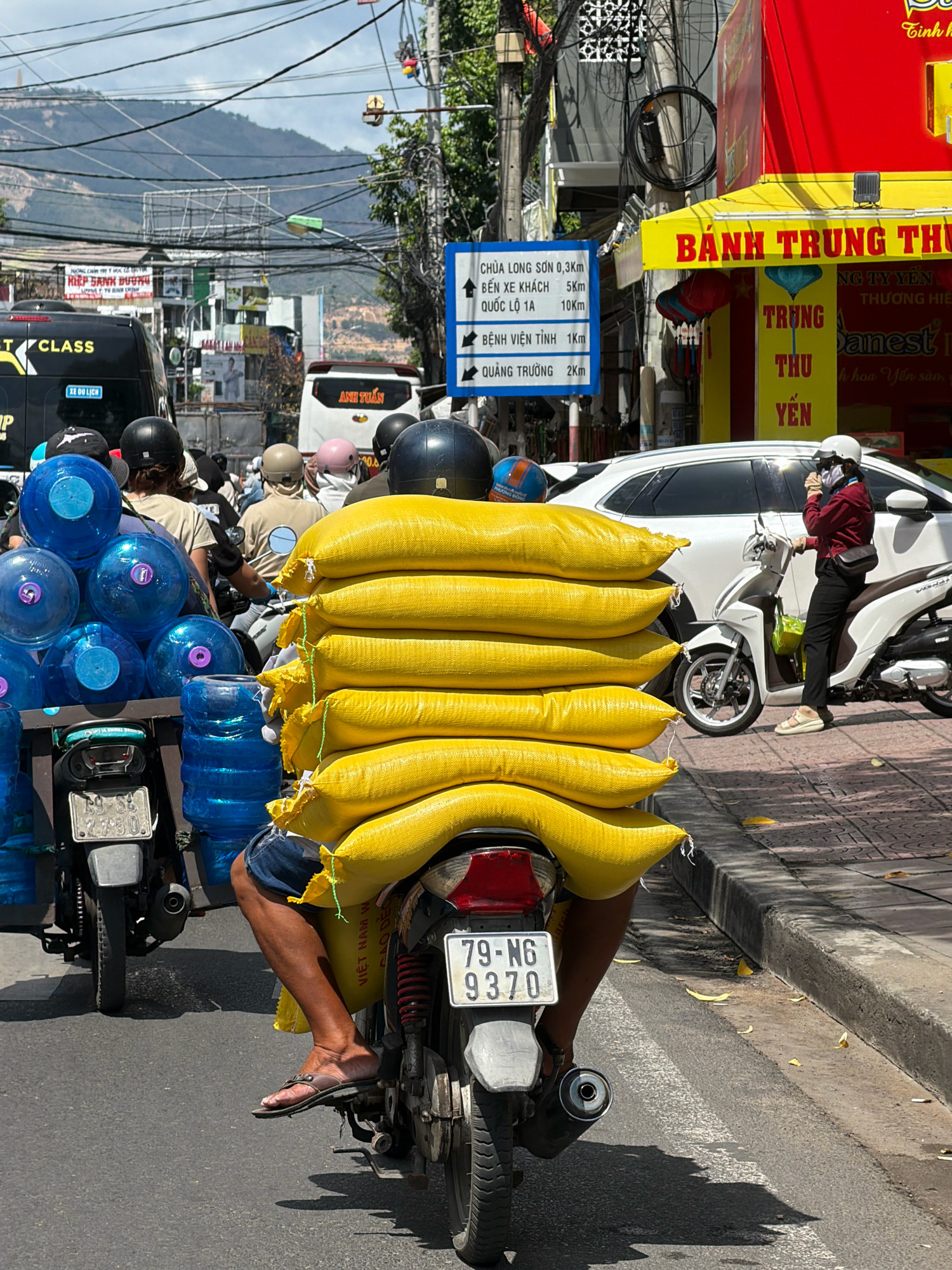 The image depicts a street scene in Nha Trang, Vietnam, likely during daylight hours. In the foreground, a person is riding a motorcycle laden with numerous large, yellow sacks stacked vertically behind the rider. The sacks appear to be made of a woven material and are tied together. To the left of the motorcycle, a rack holds a stack of large blue water bottles. The motorcycle's license plate is visible, displaying the characters "79-N6 9370". The rider is wearing shorts and flip-flops, and their legs are visible.
In the background, the street is lined with buildings, some with signage. One prominent building has a red facade with yellow trim and text that appears to be in Vietnamese. Traffic is present, with other vehicles, including buses, visible on the road. A traffic sign with directional arrows and Vietnamese text indicating distances to various locations (e.g., "CHÙA LONG SƠN 0,3KM", "QUẢNG TRƯỜNG 2KM") is positioned on the right side of the road. Overhead, a complex network of electrical wires crisscrosses the sky, a common sight in Vietnamese cities. The environment suggests an urban setting with moderate traffic and commercial activity. The presence of mountains in the far distance indicates the proximity of the city to a more mountainous region.