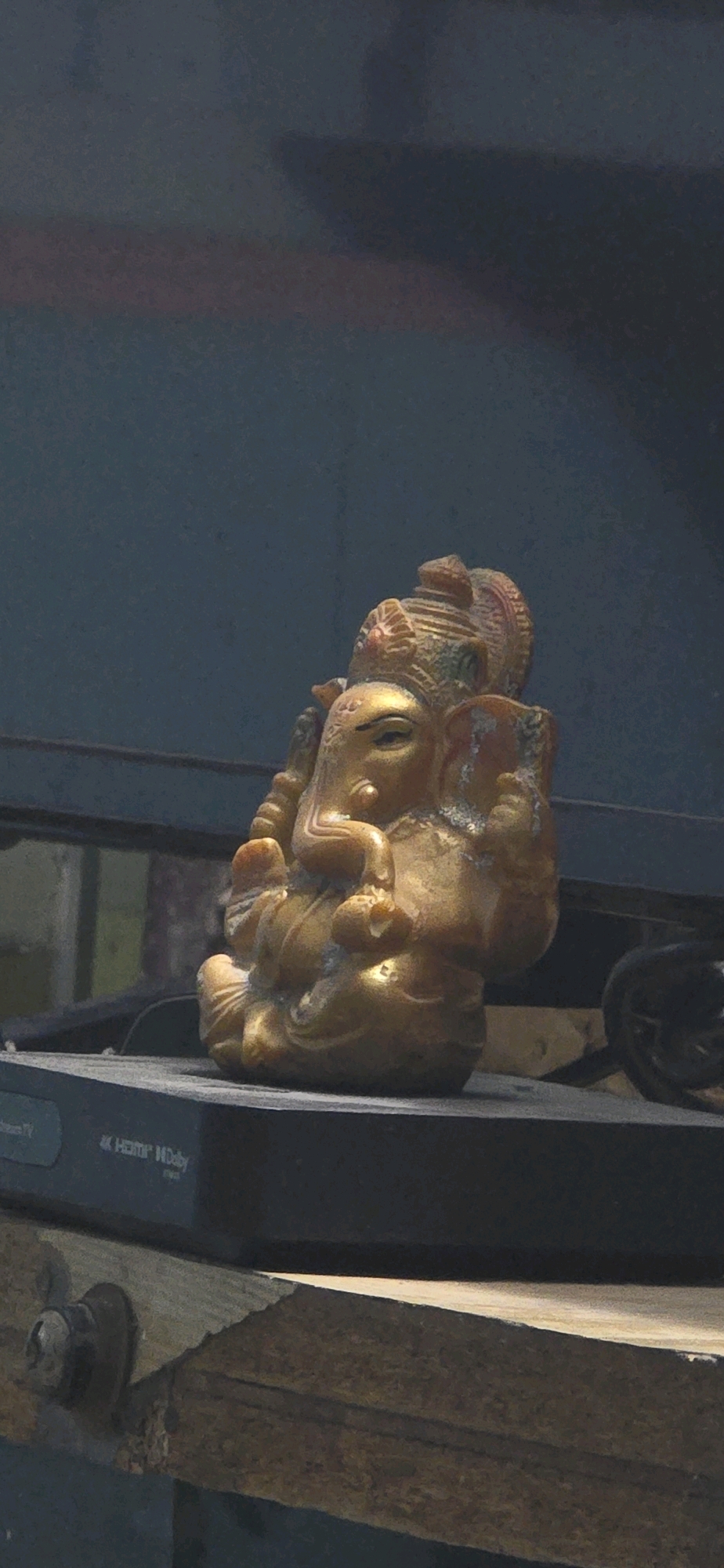 A golden figurine of the Hindu deity Ganesha sits on a dark electronic device. The figurine is ornate with visible details on its crown, face, and limbs. The electronic device appears to be a set-top box, with labels indicating "4K HDMI Dolby". The background is a dark, textured wall. The scene is illuminated by natural light, casting soft shadows. The image was captured in Kolar Gold Fields, India.