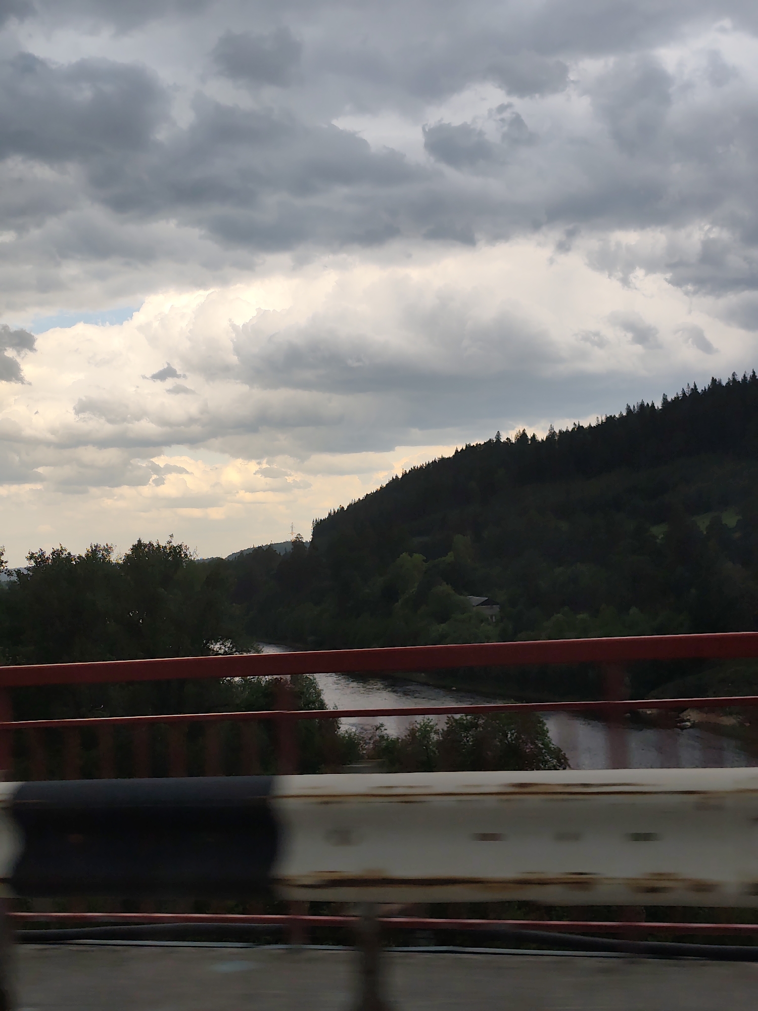 A view of a river valley from a bridge in Chusovskoye Urban Settlement, Russia. The river, reflecting the sky, is framed by dense green vegetation, primarily trees, on the left and a heavily forested hillside on the right. The sky is overcast with various cloud formations. The bridge structure includes a red painted rail and a weathered guardrail with alternating black and white sections. The photograph appears to have been taken from a moving vehicle.