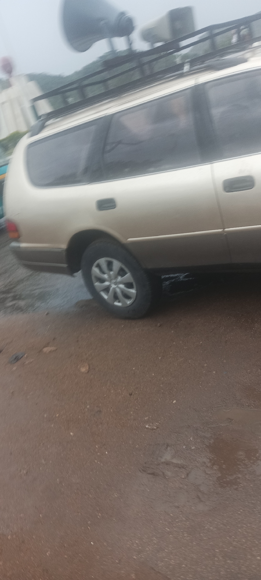 The image shows the side of a gold minivan with a roof rack and two loudspeakers mounted on top. The minivan is parked on a wet, paved surface. The location is the city of Kuru Dungus, Nigeria.