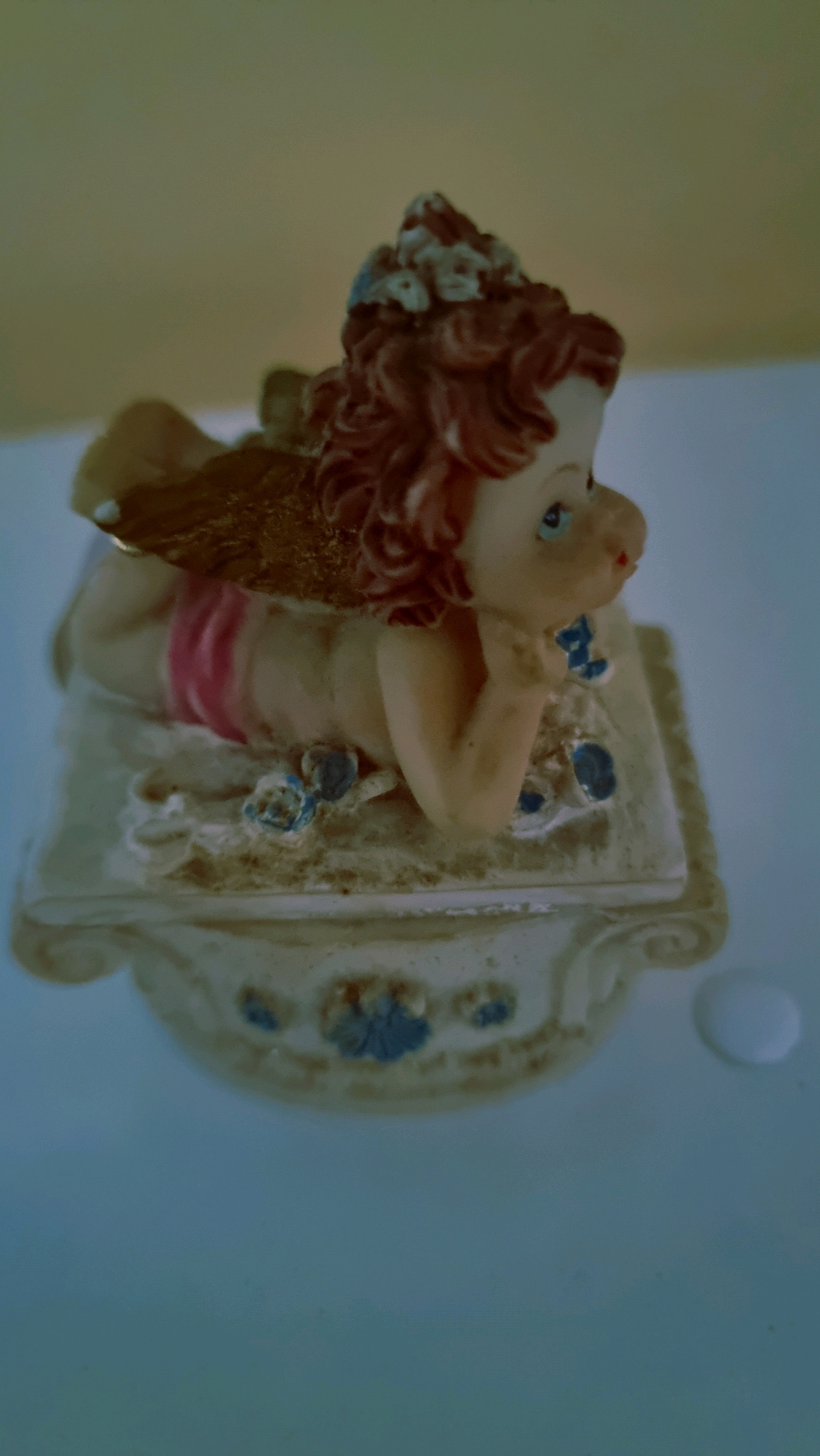 A close-up, slightly out-of-focus, low-angle shot shows a ceramic figurine of a cherub lying on its stomach on a decorative pedestal. The cherub has reddish-brown curly hair, blue eyes, and its left hand is propped under its chin, with its right arm bent. It wears pink bottoms. Its golden wings are spread behind it. The pedestal is white with decorative blue floral accents and scrollwork. The background is a blurred yellowish-beige. A small white droplet, possibly water, is visible to the right of the pedestal. The scene does not provide any specific information about its location in Sant Joan Les Fonts, Spain.