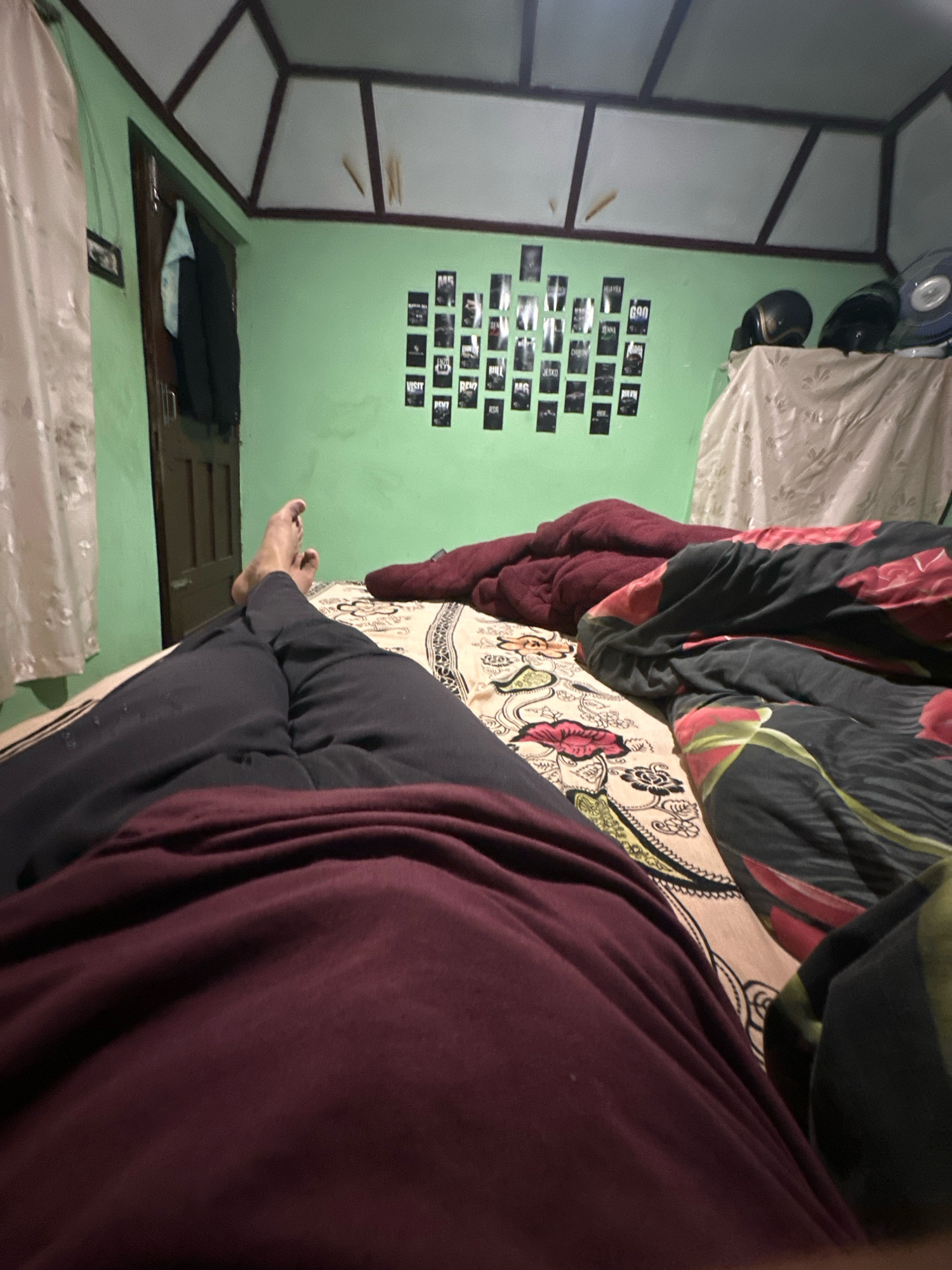An interior shot captures a bedroom from a first-person perspective, likely taken from a bed. The floor is partially visible, covered by a dark red blanket in the foreground. To the left, a person's legs, clad in dark pants and a maroon long-sleeved shirt, are extended. The bedsheets have a complex pattern in shades of cream, brown, and pink, with a floral motif. To the right, a black blanket adorned with a large floral print featuring red and green petals is draped over the bed.
The room's walls are painted a pale green, and the ceiling features a geometric design with dark brown trim. On the left wall, near a door, a framed picture is hung. A light blue towel and a dark jacket hang from the door. On the back wall, a grid of small black-and-white posters or photographs is displayed, featuring text and images. Above these, on a table or shelf covered by a light brown patterned cloth, three motorcycle helmets are placed: two black and one blue. A fan is visible in the upper right corner. The lighting in the room appears dim.