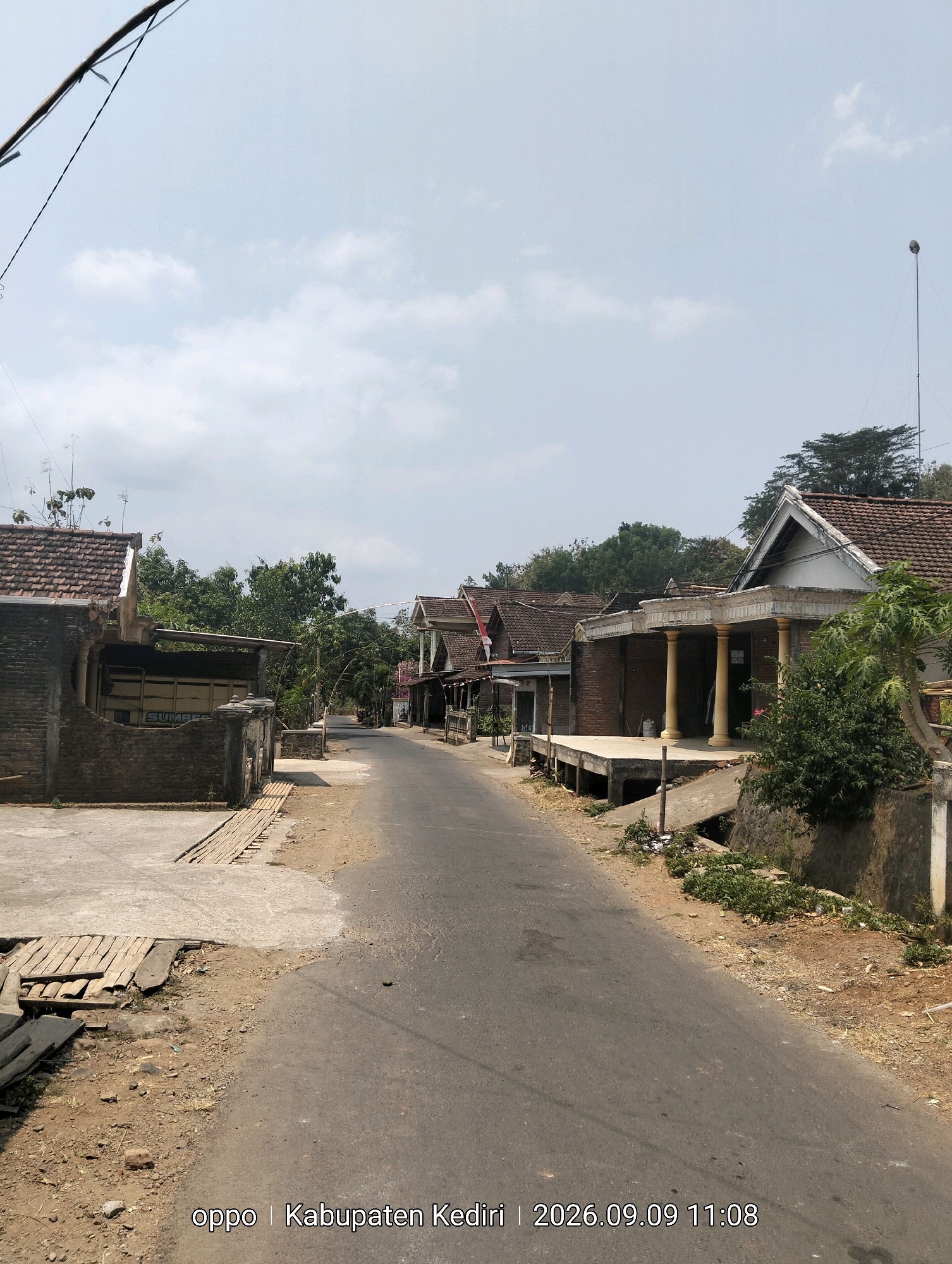 The image shows a residential street in Pagung, Indonesia, on a bright, overcast day. The street is paved and appears to be a two-lane road with no visible lane markings. On the left side of the street, there is a property with a brick wall and a covered carport. Further down the street on the left, several houses with tile roofs are visible, interspersed with trees. On the right side of the street, more houses with tile roofs are seen, some featuring porches with columns. The houses are of a traditional Indonesian architectural style. The surrounding vegetation consists of various trees and bushes, indicating a rural or semi-rural setting. The ground along the side of the road is dirt and gravel. A utility pole with wires is visible in the upper left portion of the image, and a lamppost is present in the upper right. The bottom of the image contains a watermark with "oppo | Kabupaten Kediri | 2026.09.09 11:08". There are no people or vehicles visible in the scene.