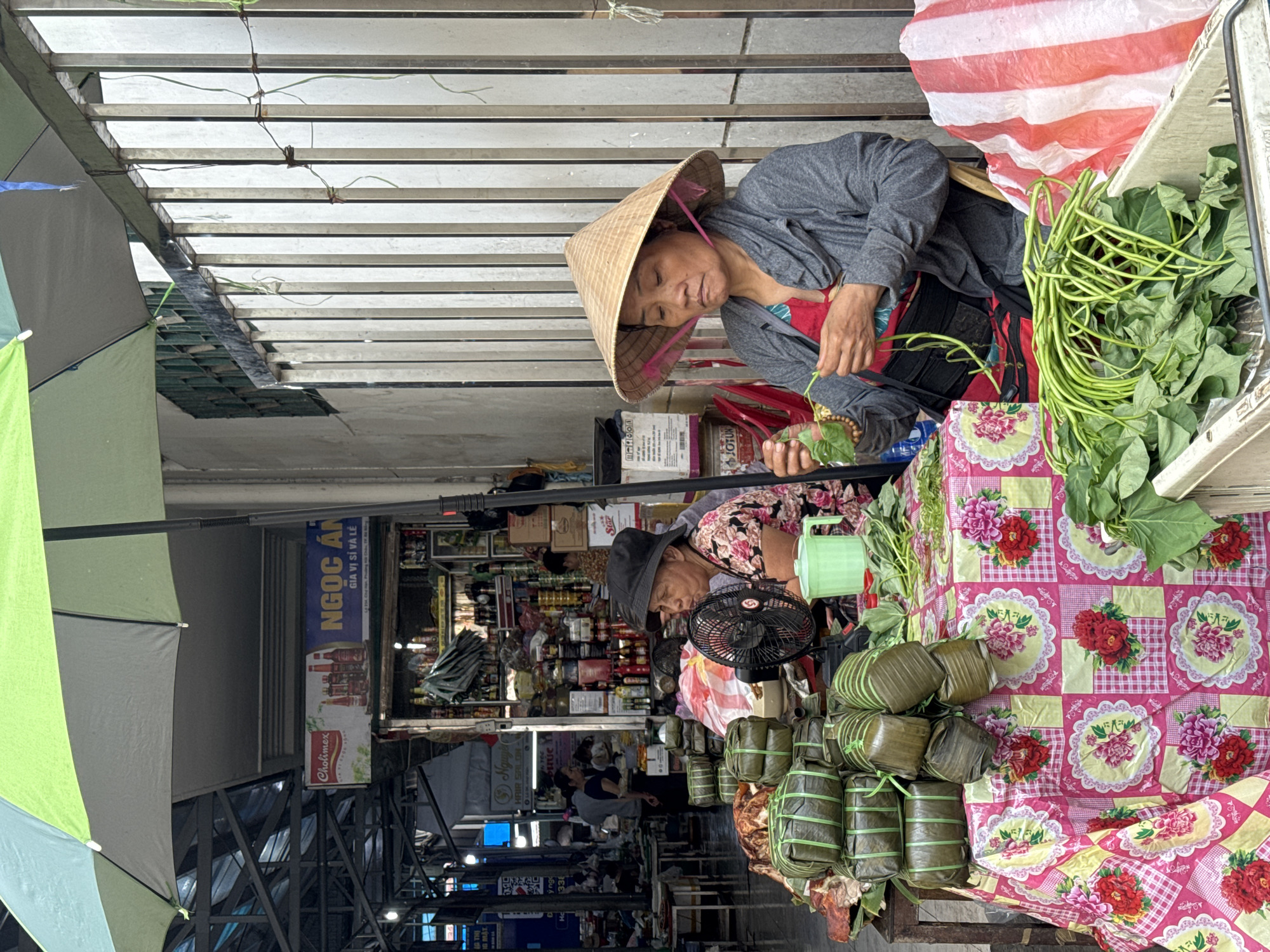 Two Vietnamese women are selling goods at a market. The woman on the top right is wearing a conical hat and a grey jacket and is sorting through a bundle of leafy greens. The other woman is wearing a floral shirt and a black hat, and a small black fan is placed in front of her. There are stacks of cylindrical food items wrapped in leaves and tied with green string on the table. The table is covered with a pink tablecloth decorated with flowers. Behind the women, a shop displays various goods on shelves. A sign with the words "NGOC AN GIA VI SI VA LE" is visible. The market is covered by a metal structure and an umbrella provides additional shade.
