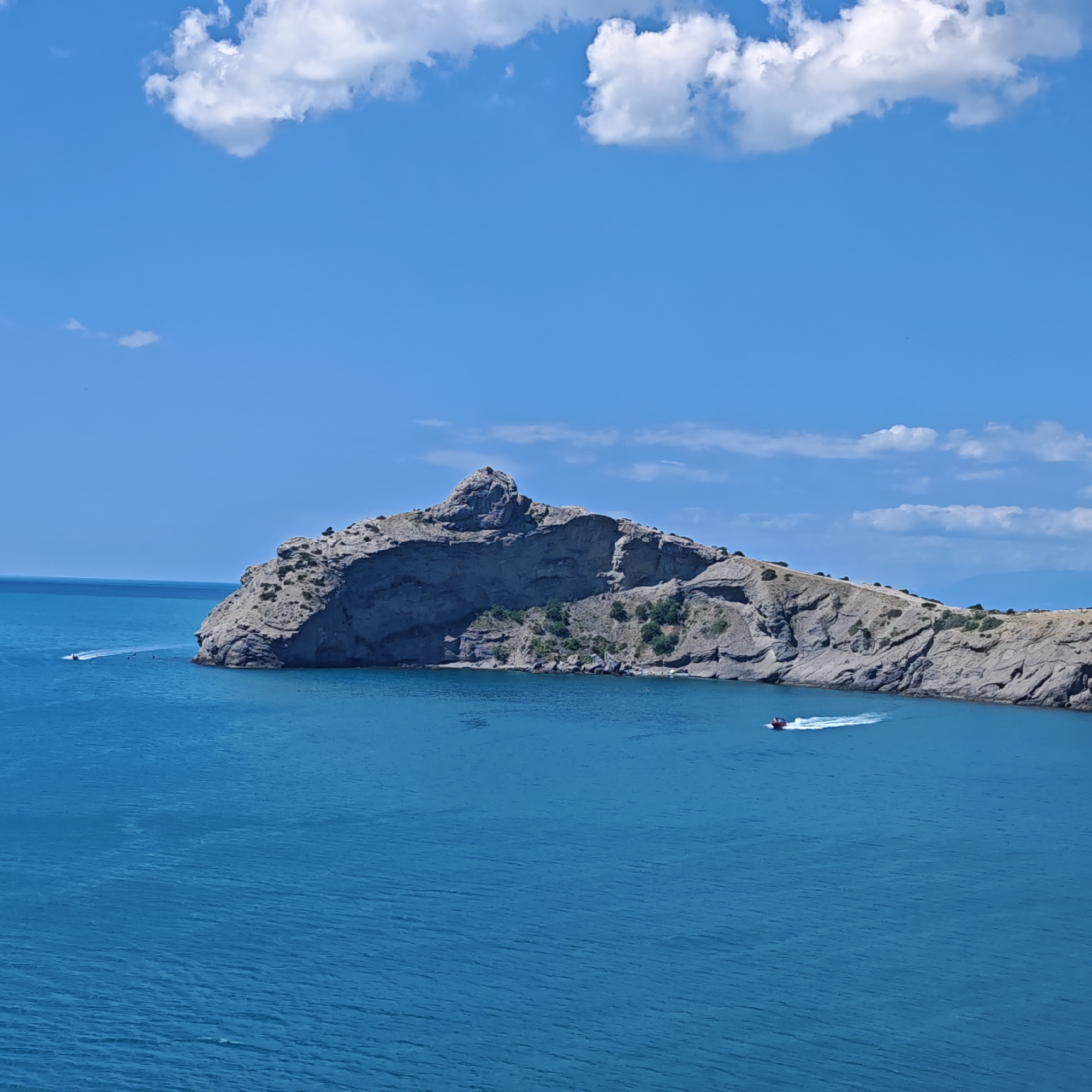 An elevated, wide-angle, full shot shows a body of bright blue water and a rocky, mountainous coastline under a clear sky with scattered white clouds. The coastline juts out into the water, with a prominent, jagged rock formation at its peak. Small patches of green vegetation are visible on the slopes of the cliffs. Two red and white speedboats are visible in the water; one is farther out to sea and appears to be moving away from the viewer, while the other is closer to shore and is creating a white wake as it moves to the right. The overall scene is serene and depicts a sunny day at the seaside, likely in a coastal area near Sudak, Ukraine, given the characteristic rocky formations and azure waters.